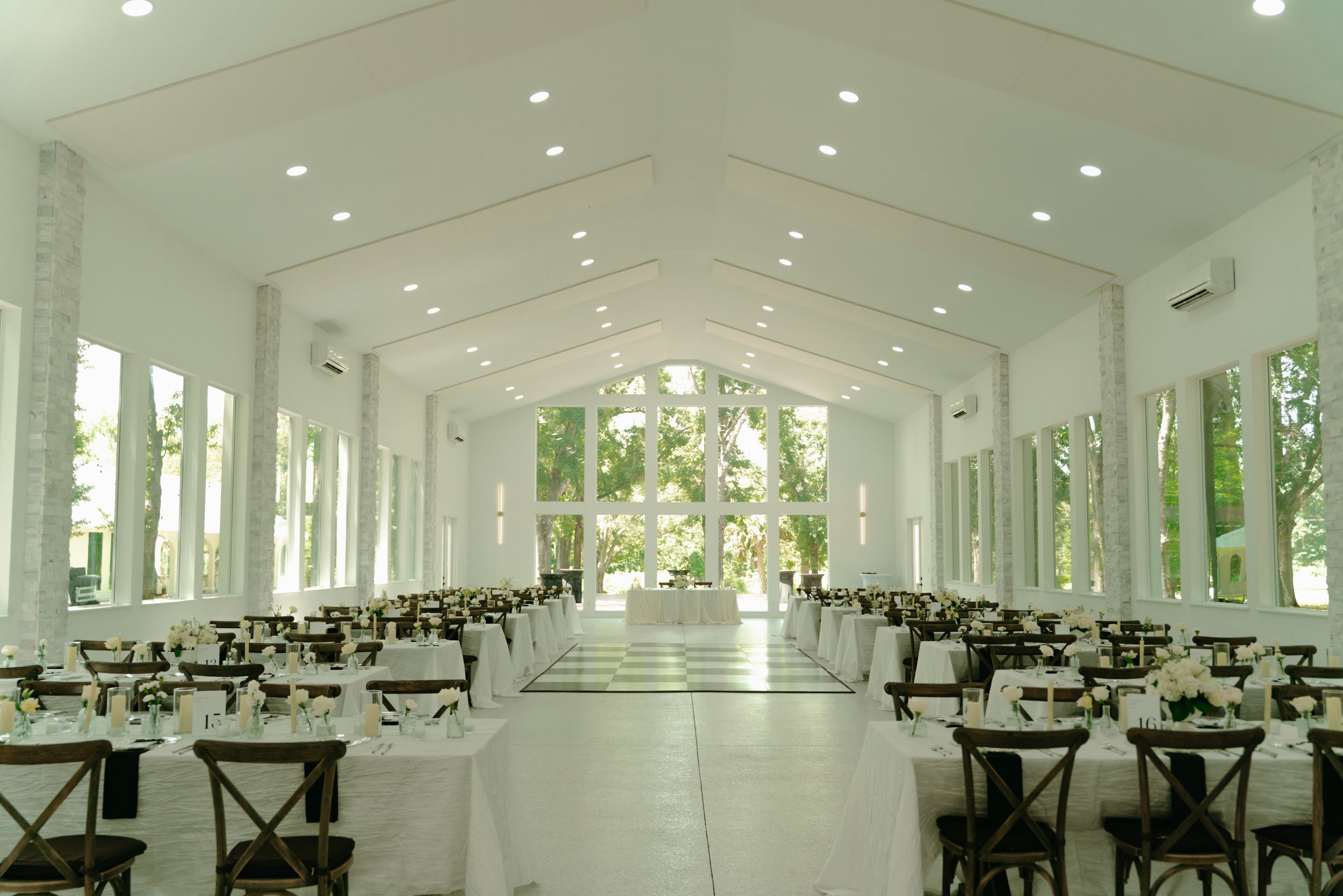 Bright white wedding reception hall with high vaulted ceilings, floor-to-ceiling windows, a checkered dance floor in the center, and long tables with white linens, wooden cross-back chairs, candles, and white floral arrangements.