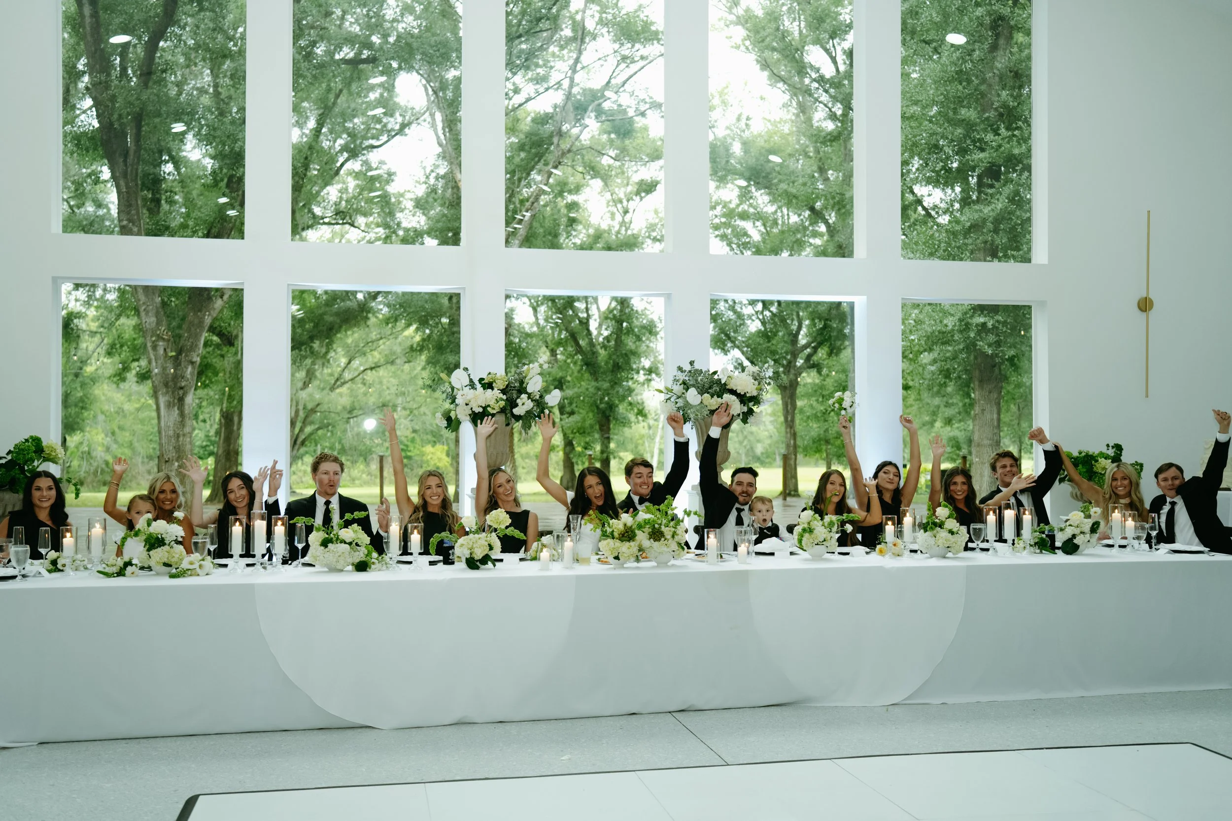 Wedding party seated at a long white head table, raising their hands and bouquets in celebration, with white floral centerpieces, candles, and large windows showing green trees behind them.