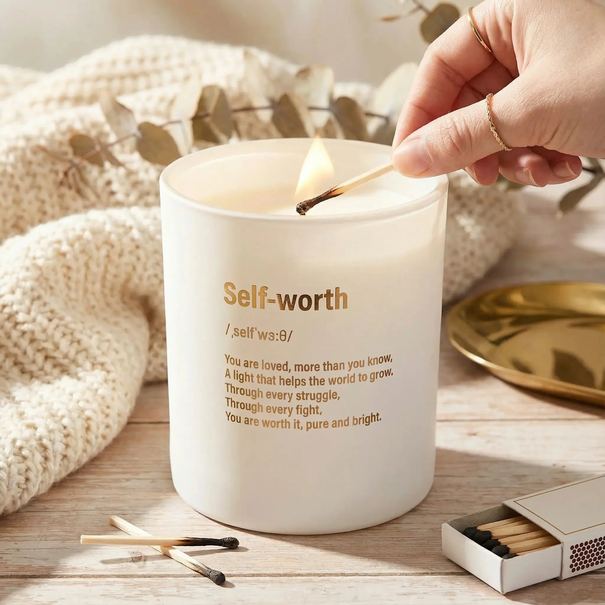 A white candle with the word 'Self-worth' and a definition printed on it, with a person lighting a matchstick to the candle. Surrounding items include a beige knitted cloth, a small box of matches, and a gold dish on a wooden surface.