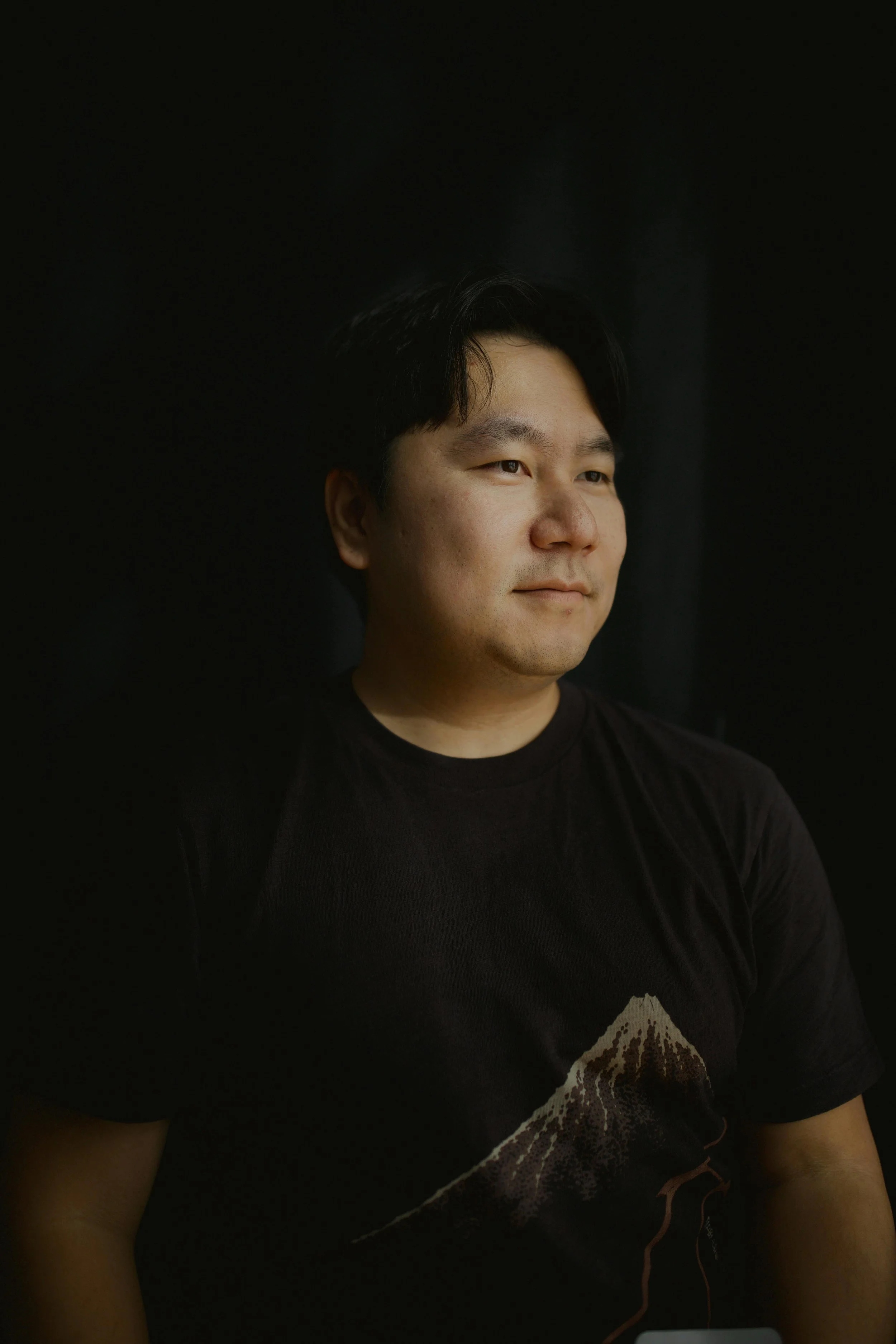 Portrait of a young man with black hair, wearing a black t-shirt with a mountain graphic, against a dark background.