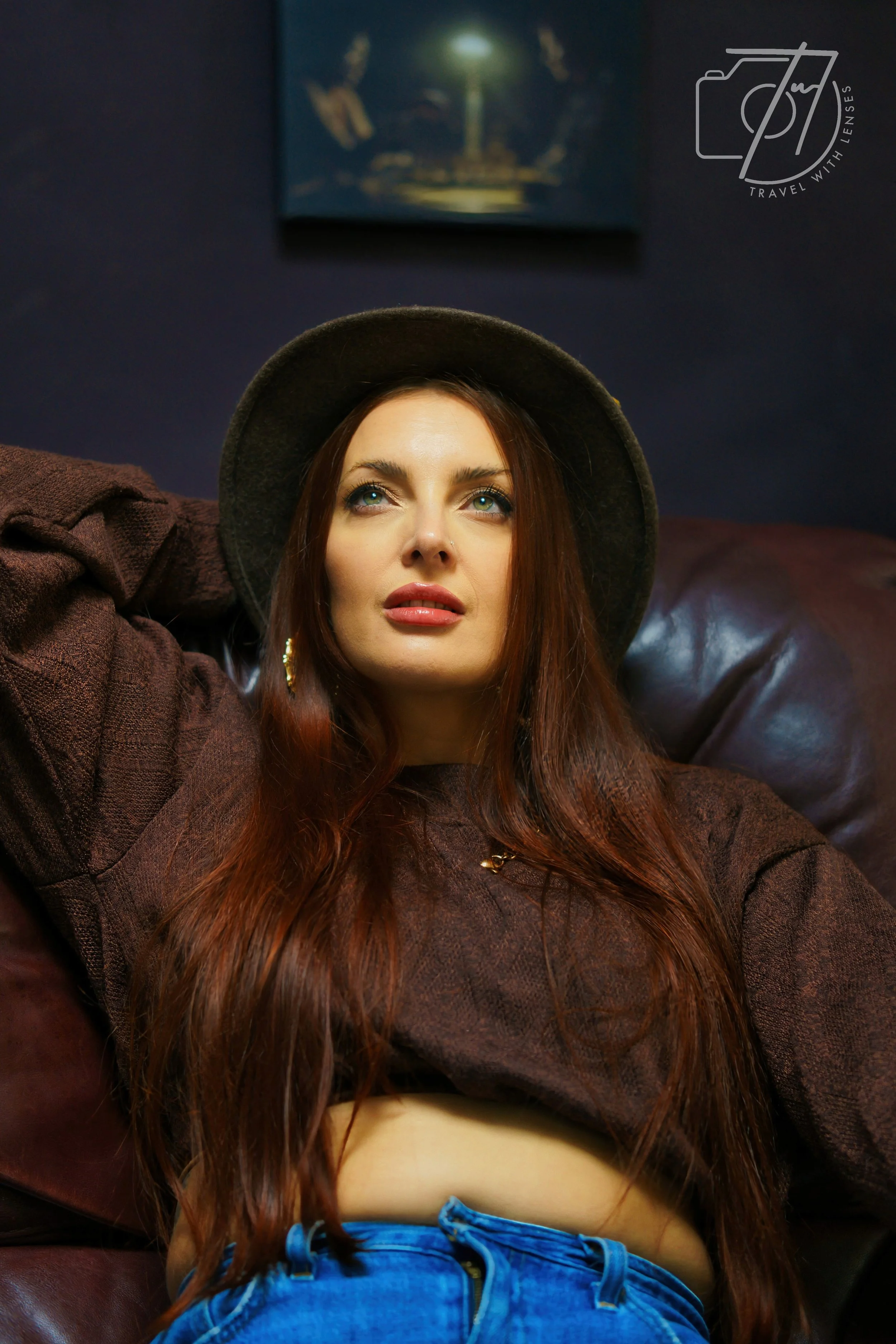 A young woman with long red hair wearing a black hat, a brown shirt, and blue jeans, sitting on a leather couch in a dimly lit room with dark walls and a painting in the background.