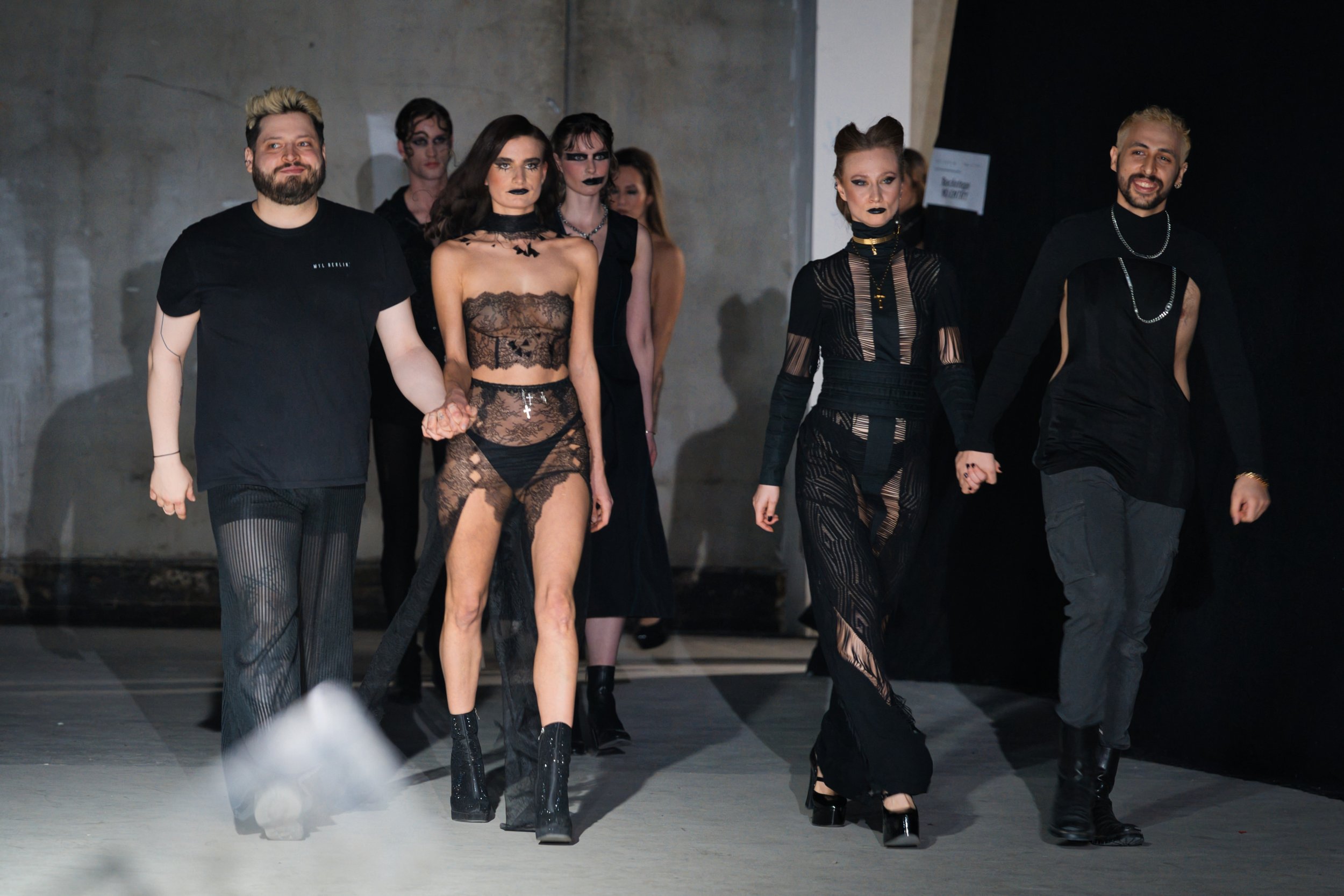 Models walking together at a fashion show, dressed in black, edgy, and gothic-inspired outfits, with some wearing lace and sheer fabrics.
