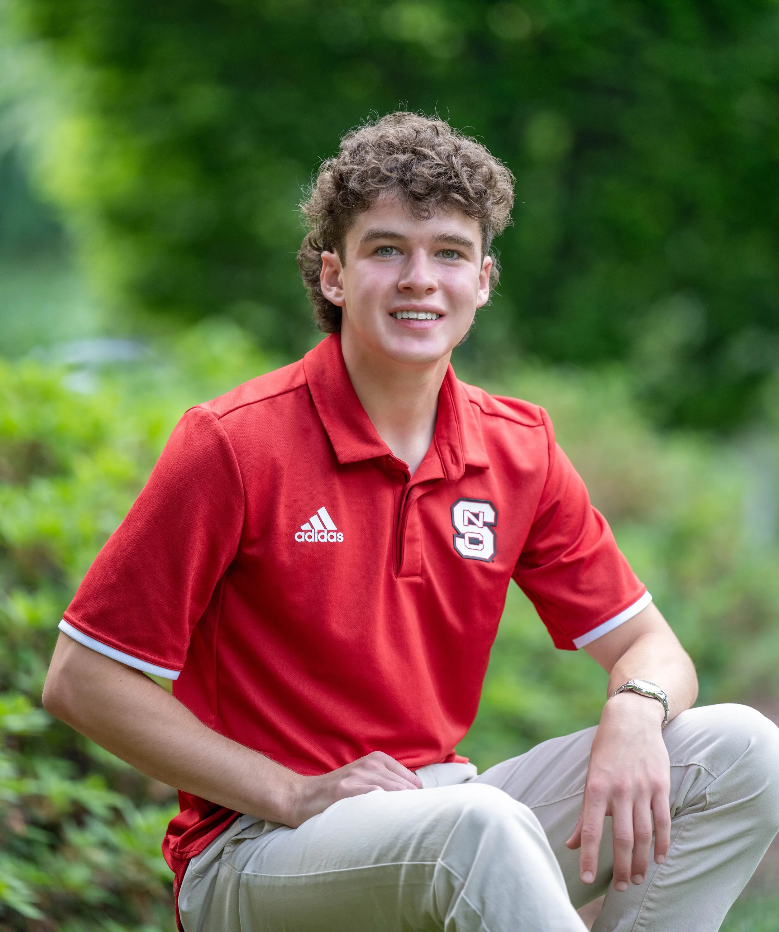 Young man wearing a red NC State polo shirt, sitting outdoors with a green, blurred background.