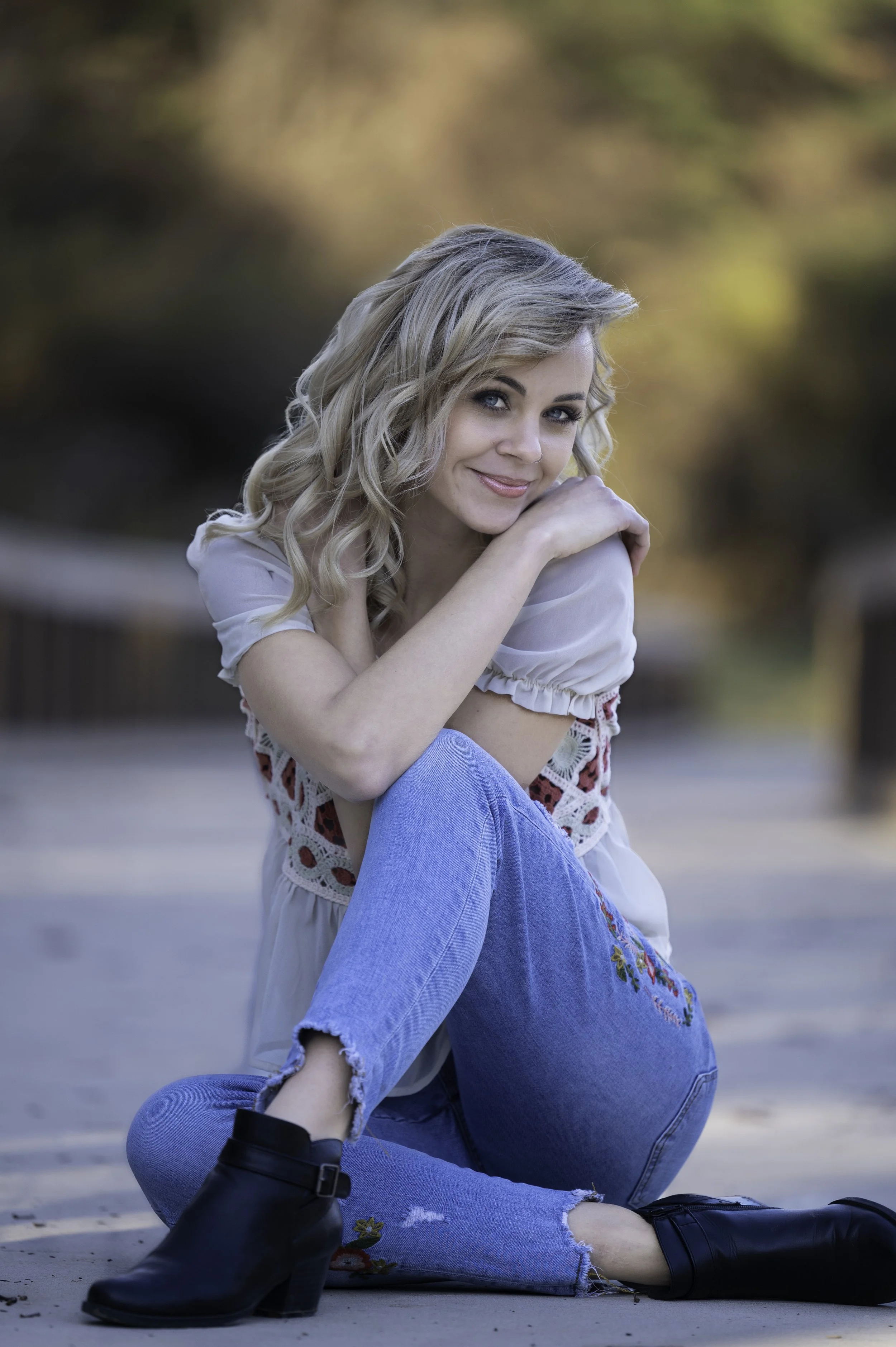 Woman with curly blond hair sitting in an outdoor setting, wearing a white blouse and blue jeans