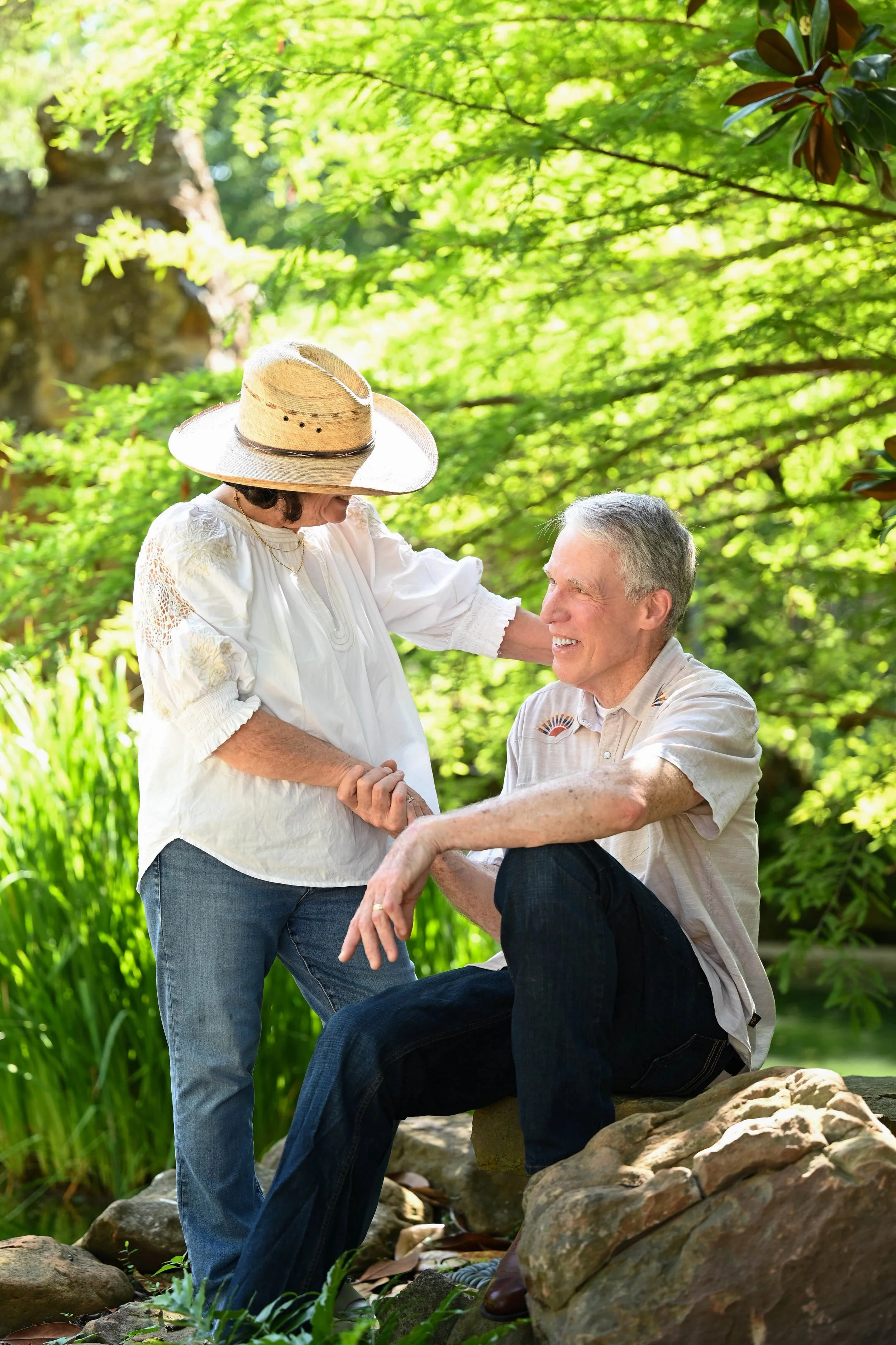 An elderly man sitting on a rock and an elderly woman standing next to him, smiling and holding hands in a lush green garden with sunlight filtering through leaves.