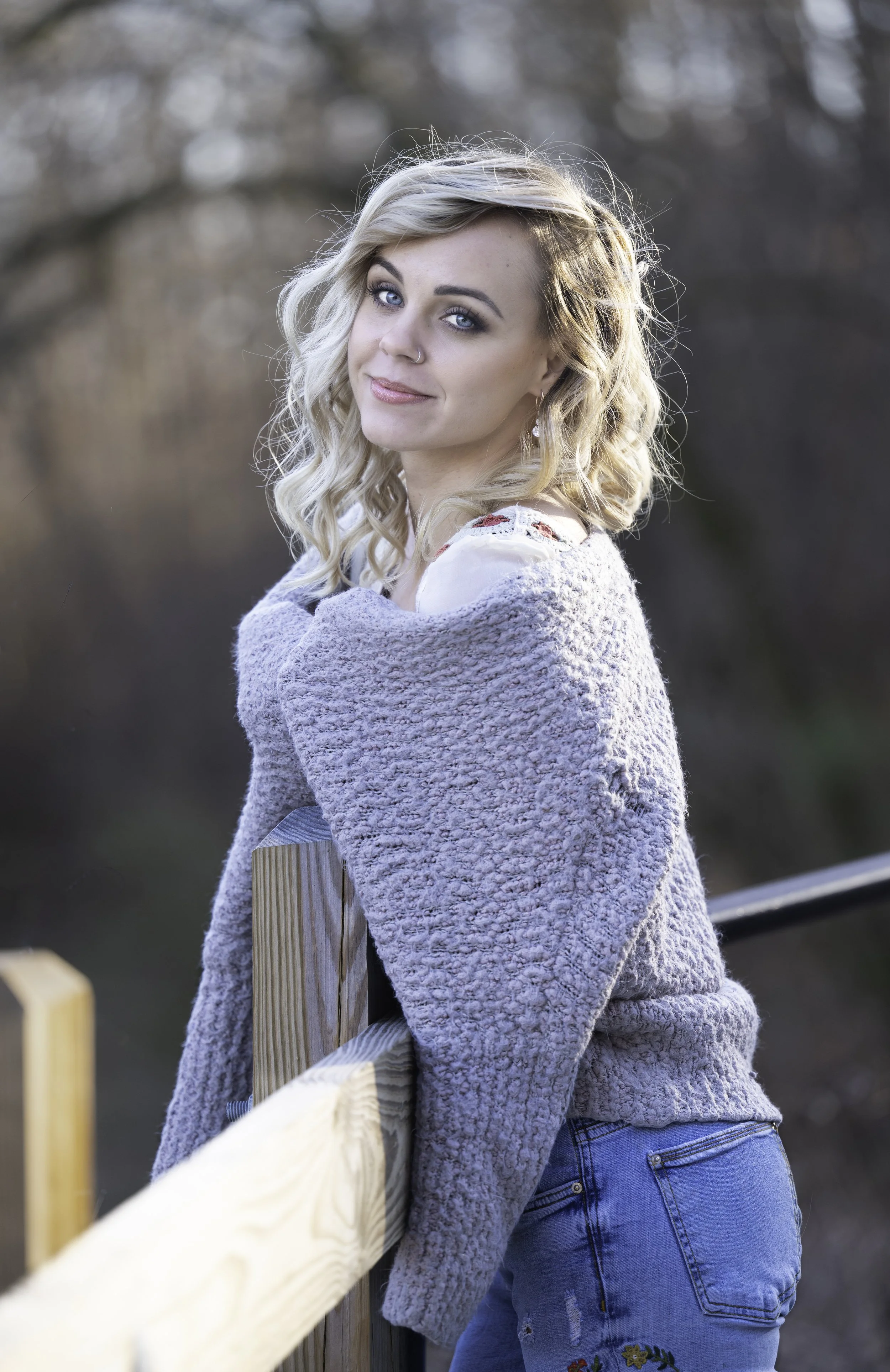 Woman with blonde hair leaning on wooden railing, wearing a gray sweater and blue jeans, outdoors.