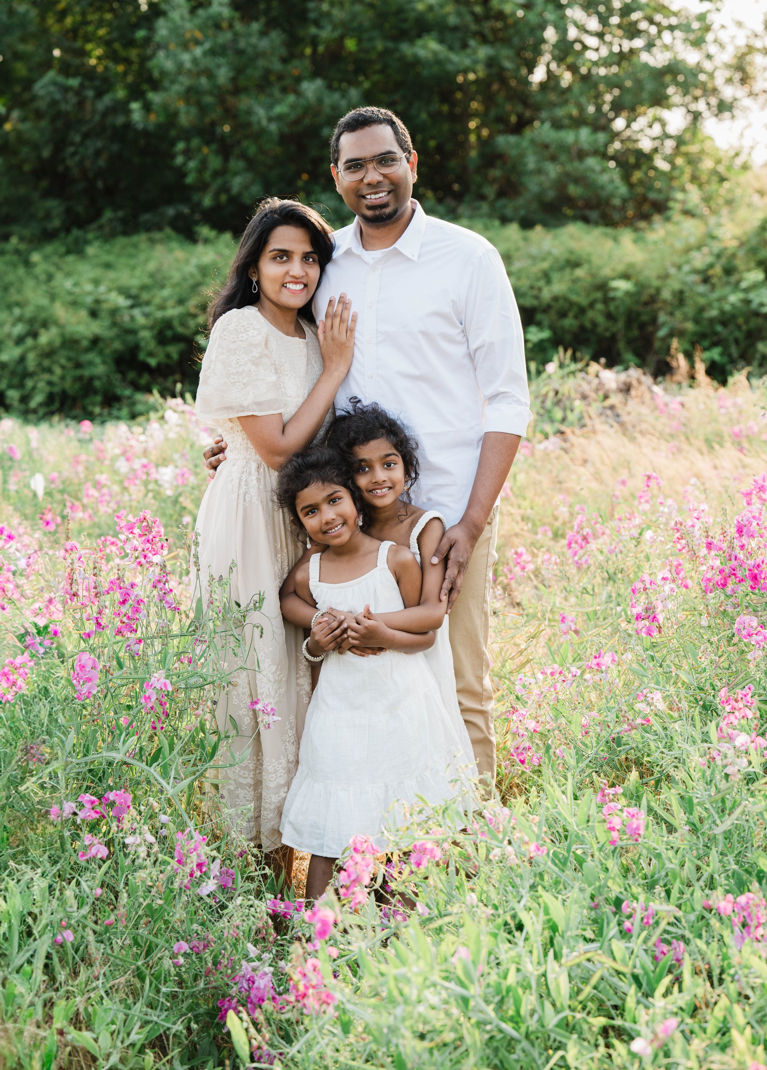 Family of four standing in a field of pink and purple wildflowers, with trees in the background. The adults and children are smiling, wearing light-colored clothing.