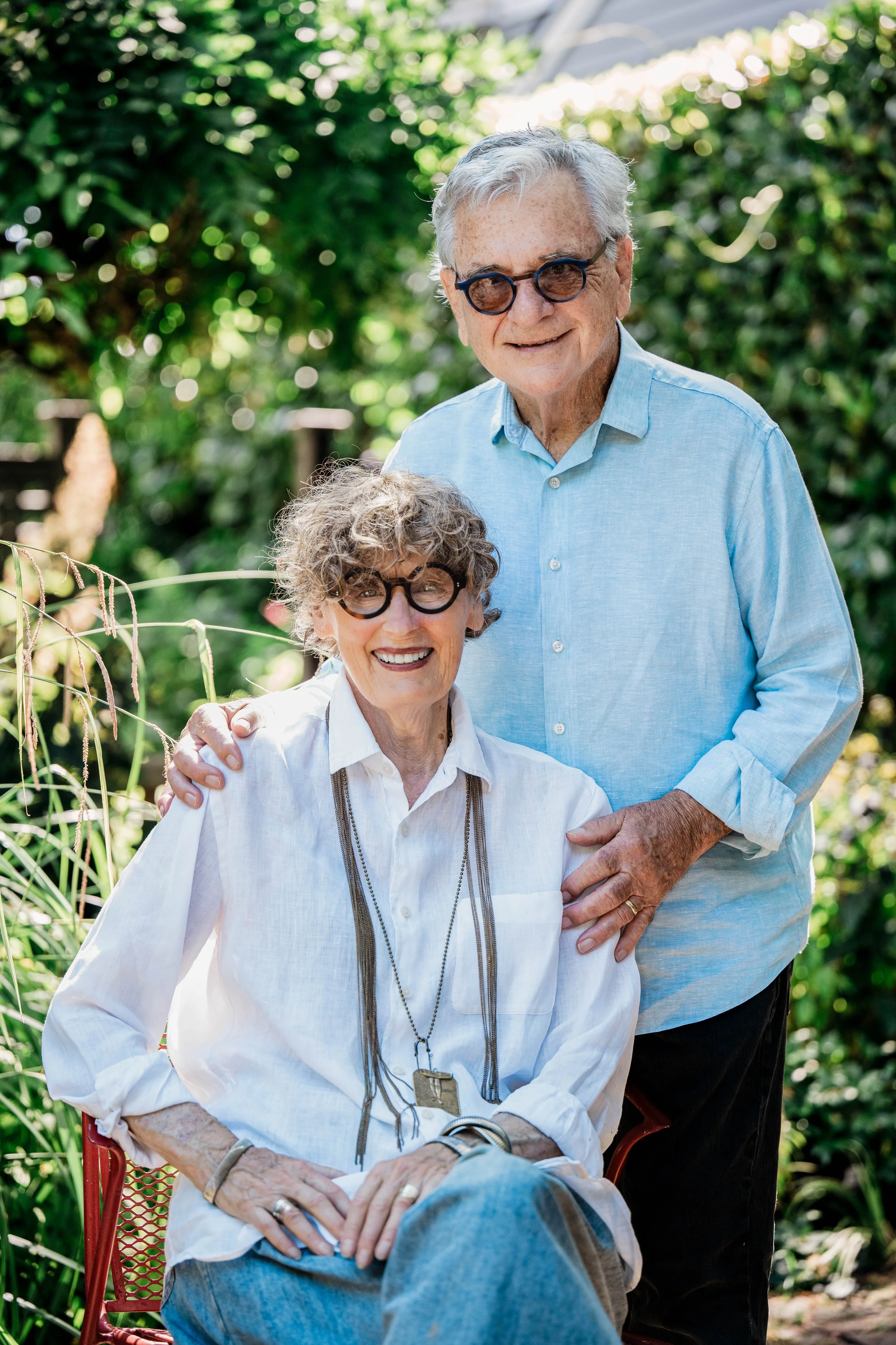 An elderly couple smiling in a garden, the woman seated and the man standing behind her with a hand on her shoulder. They are both wearing glasses and light-colored clothing.