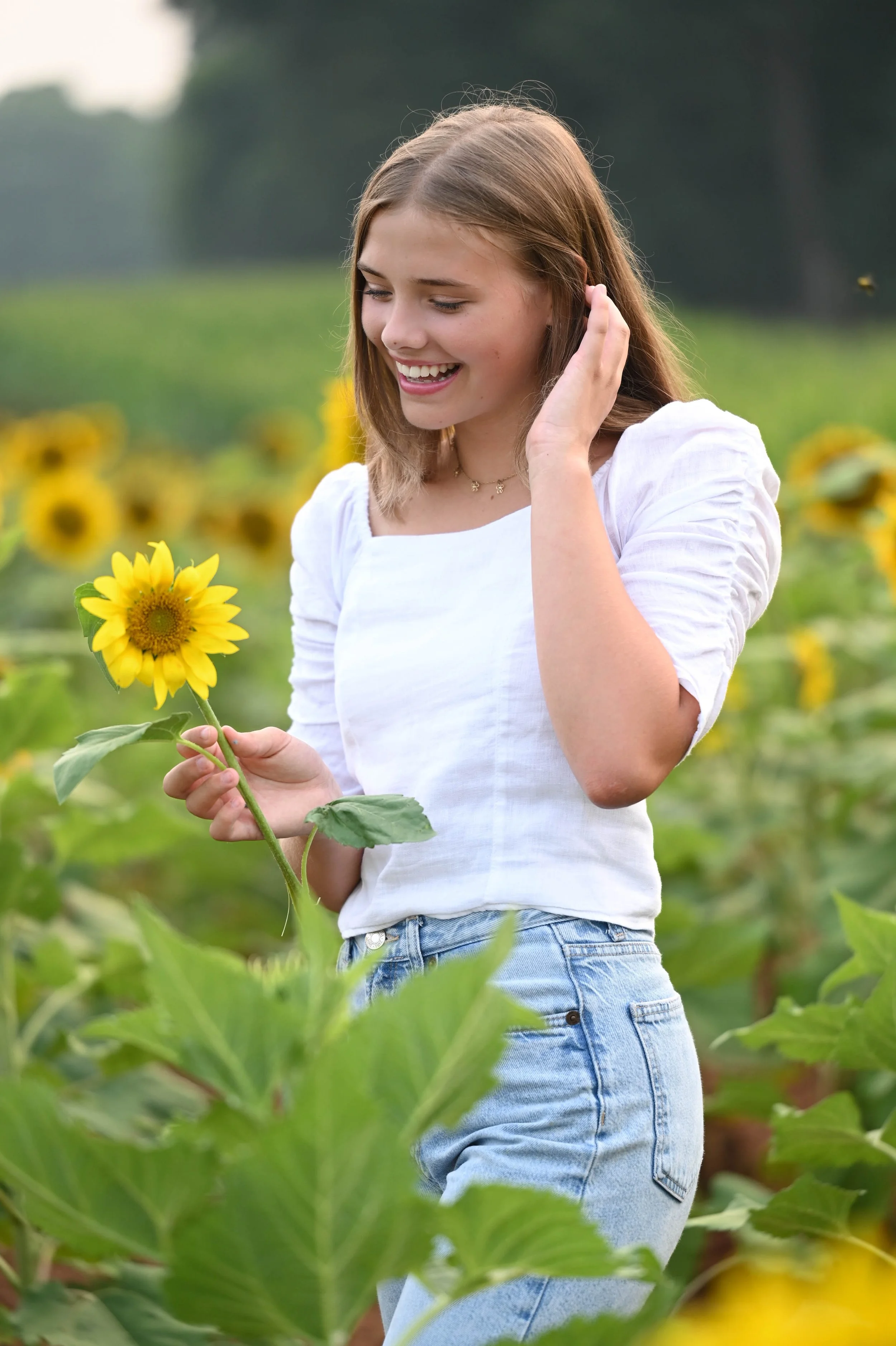 Person in white blouse and jeans holding a sunflower in a field, smiling.