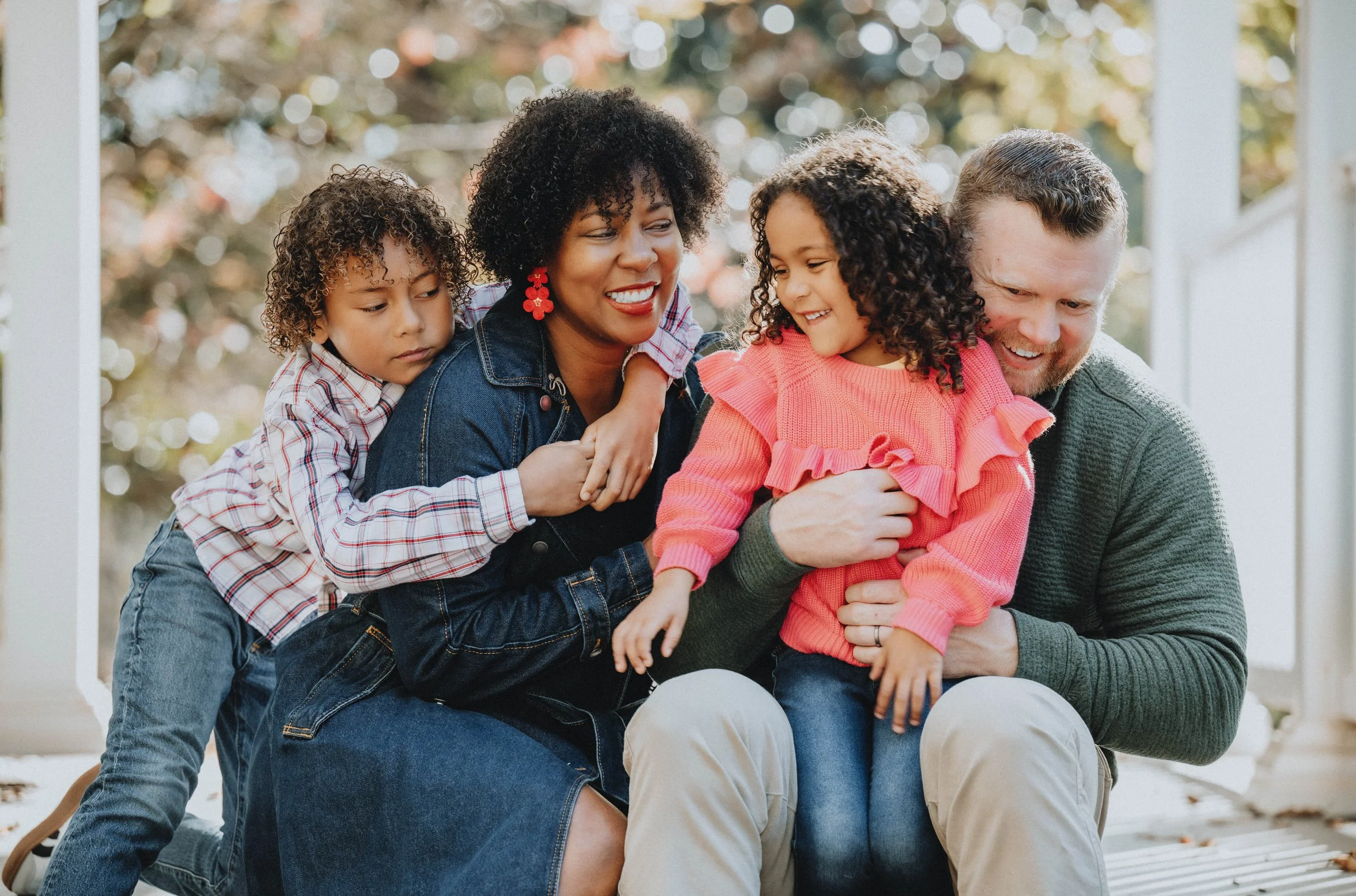 A happy multiethnic family of four enjoying time together outdoors on a porch, with two children hugging and sitting on their parents' laps.