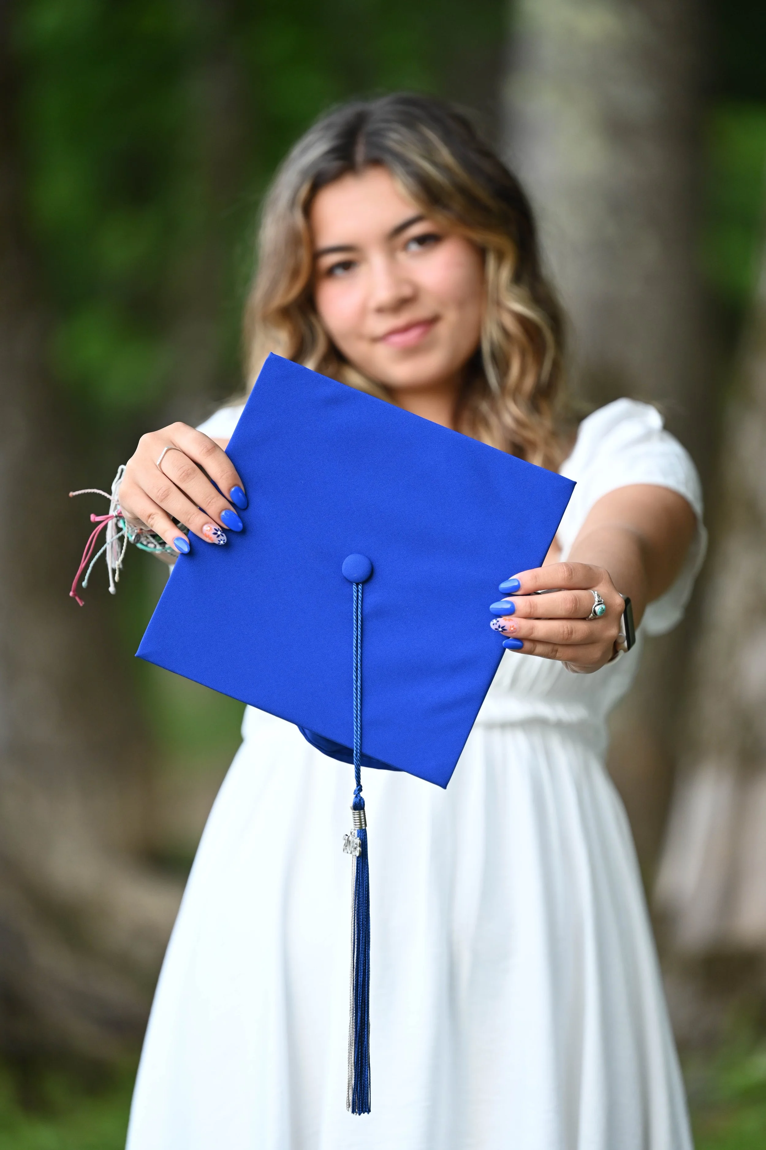Young woman in a white dress holding a blue graduation cap with a tassel in an outdoor setting.
