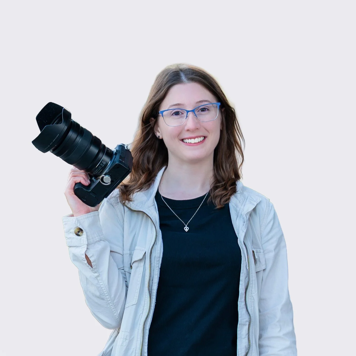 A smiling woman with blonde hair, wearing a light blue button-up shirt, holding a Nikon DSLR camera with one hand, standing against a plain light gray background.