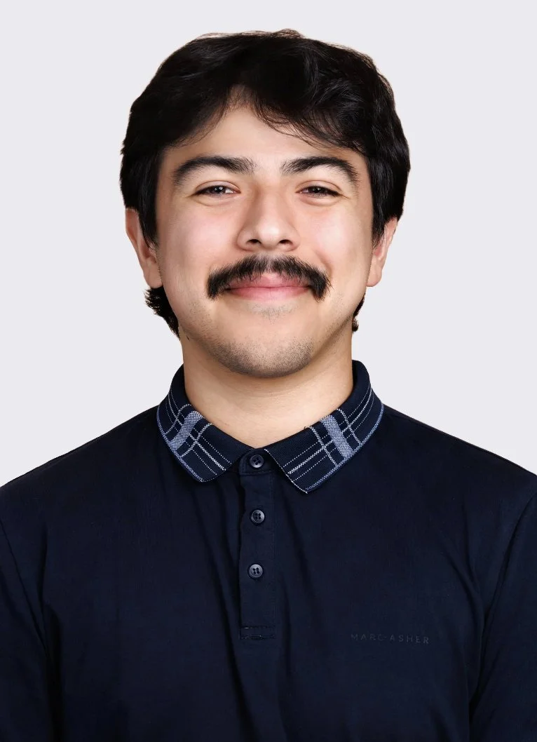 A smiling man with dark hair and a mustache, wearing a black polo shirt with a patterned collar, standing against a plain gray background.