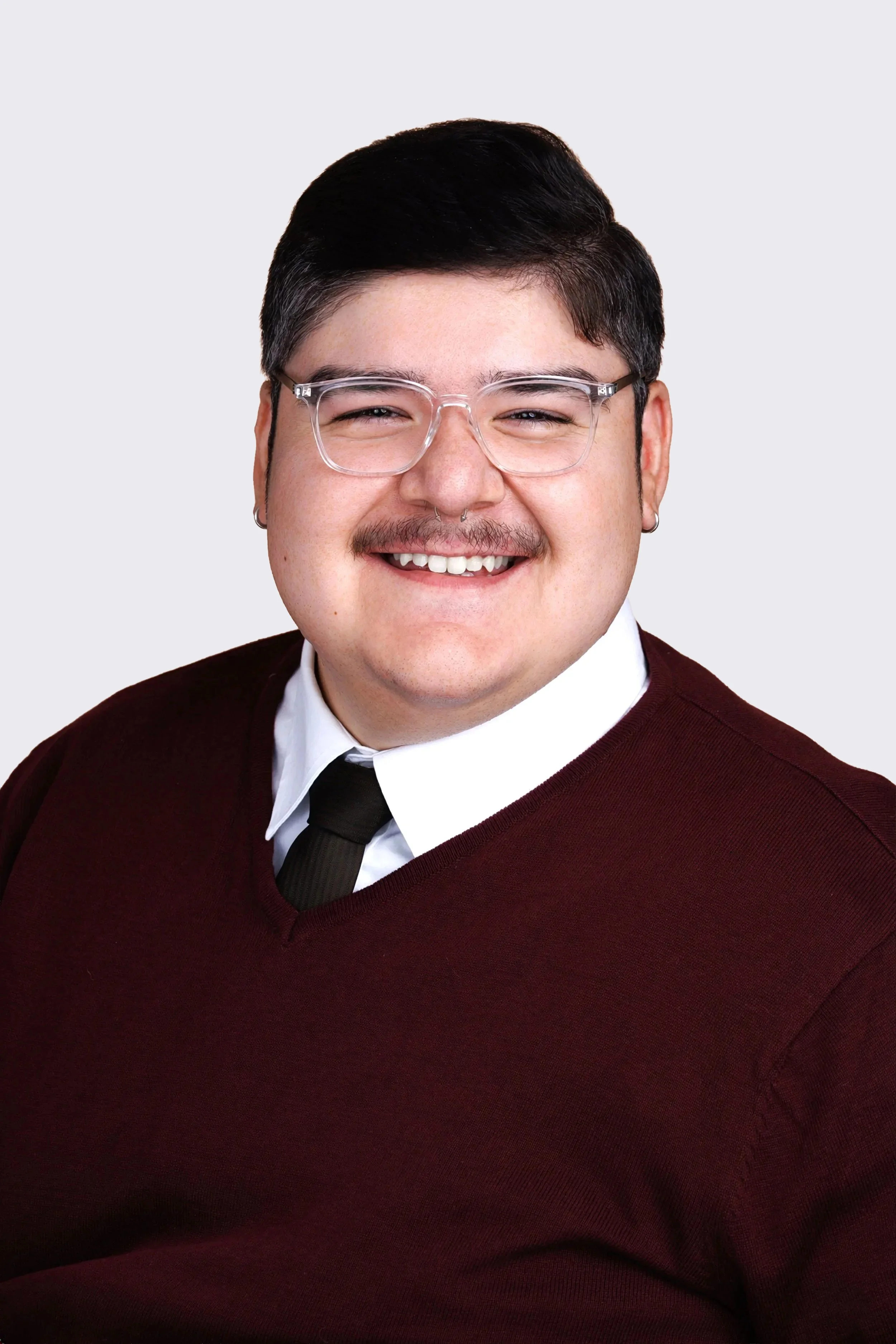 Close-up of a person with short dark hair, clear glasses, a mustache, and earrings, smiling at the camera, wearing a burgundy sweater over a white shirt with a black tie, against a plain light gray background.