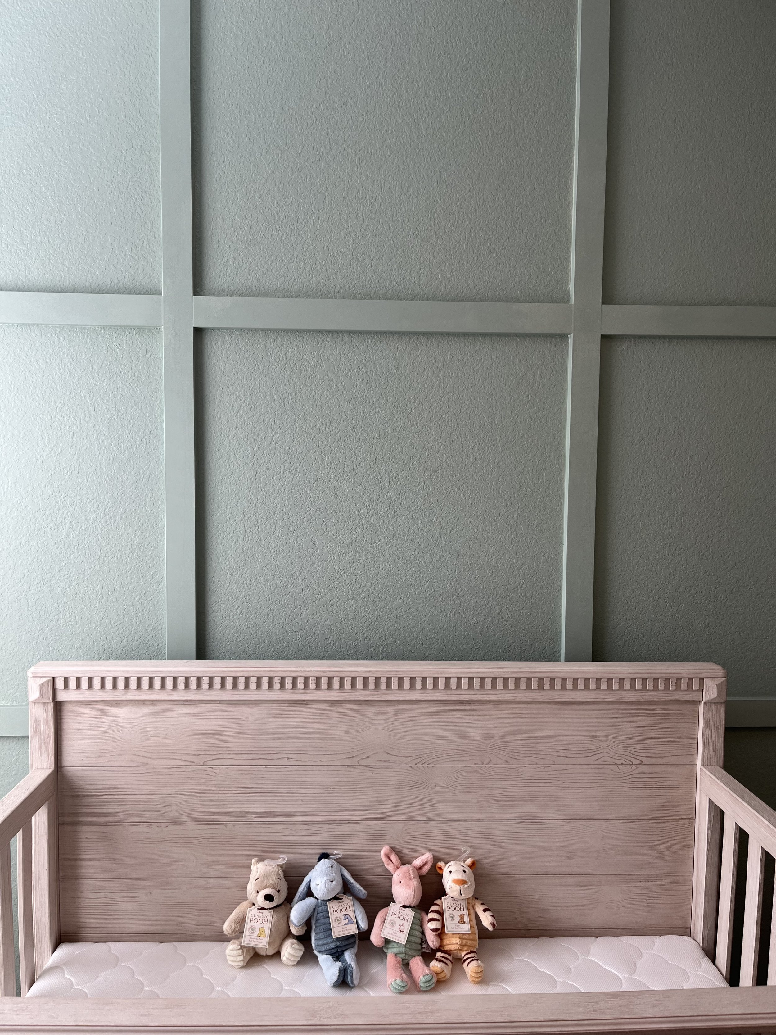 Four plush toys of Winnie the Pooh, Eeyore, Piglet, and Tigger sitting on a white mattress in a wooden crib against a plain wall.