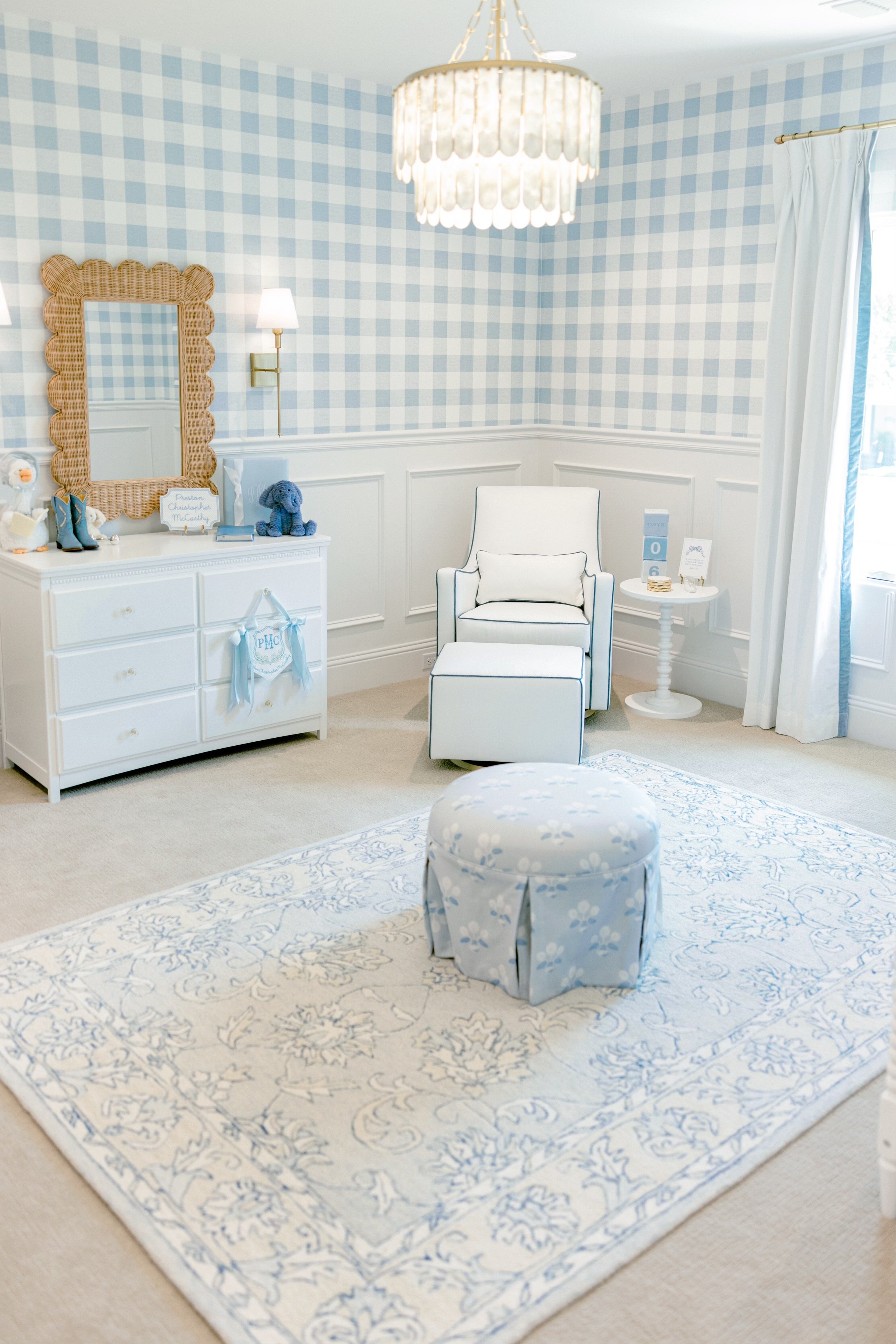 A nursery room with blue and white checkered wallpaper, white furniture, a patterned rug, a white armchair with a matching ottoman, a round side table, and decorative items including a mirror and plush toys.