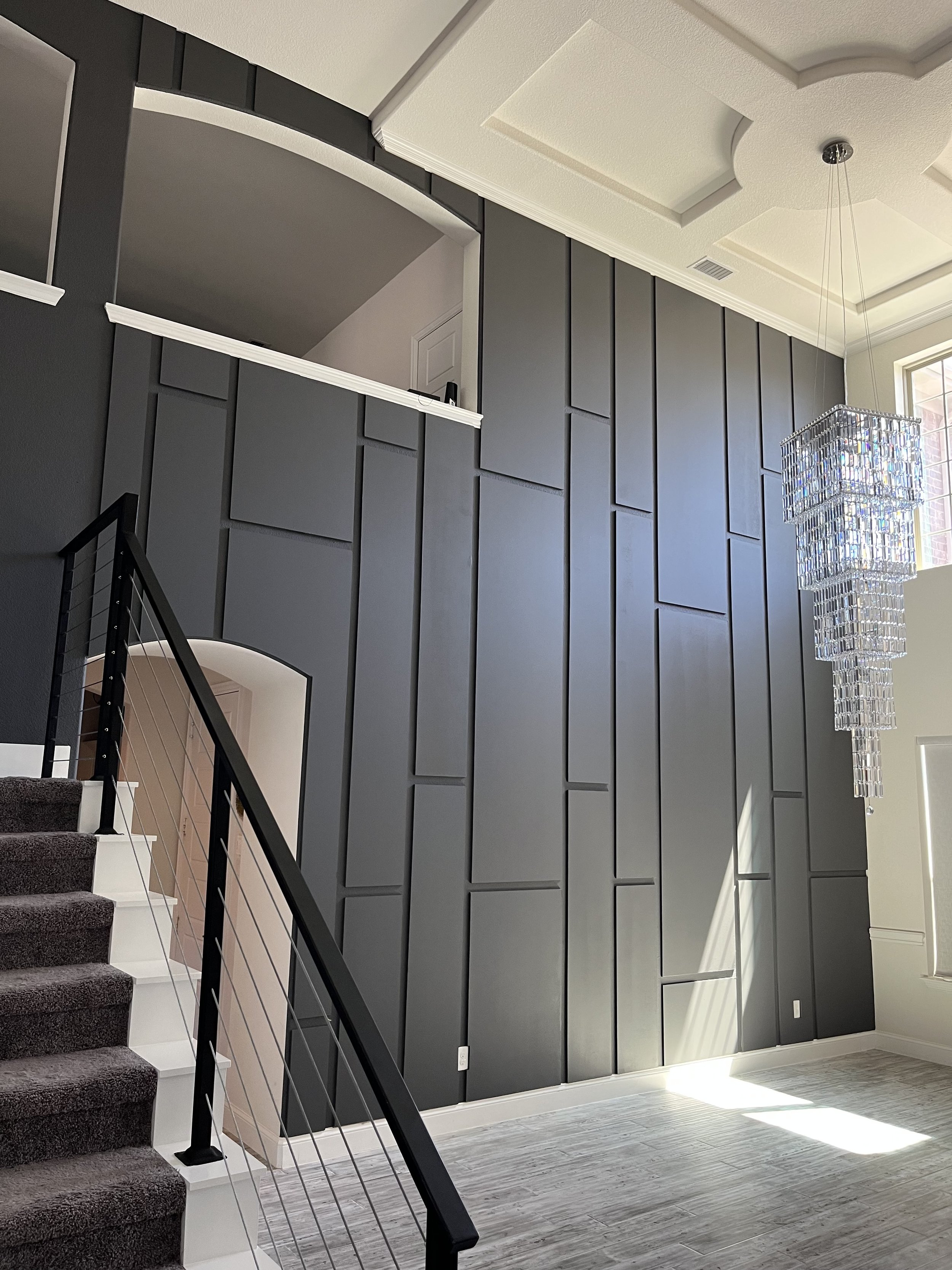Interior view of a modern staircase and a wall with dark gray panels. The staircase has black railing and white steps with dark carpeting. A window on the right side lets in natural light, and there is a large, multi-tiered chandelier hanging from th