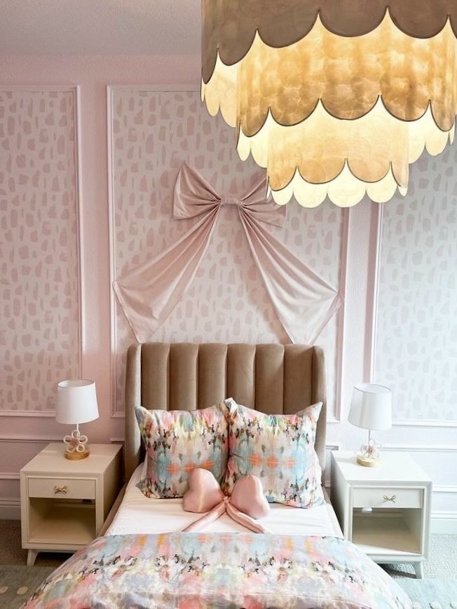 A cozy bedroom with a tall, upholstered headboard, pink and orange patterned bedspread, matching decorative pillows, and two white nightstands with lamps. There is pink fabric draped and tied into a bow on the wall behind the bed. A large, scalloped,