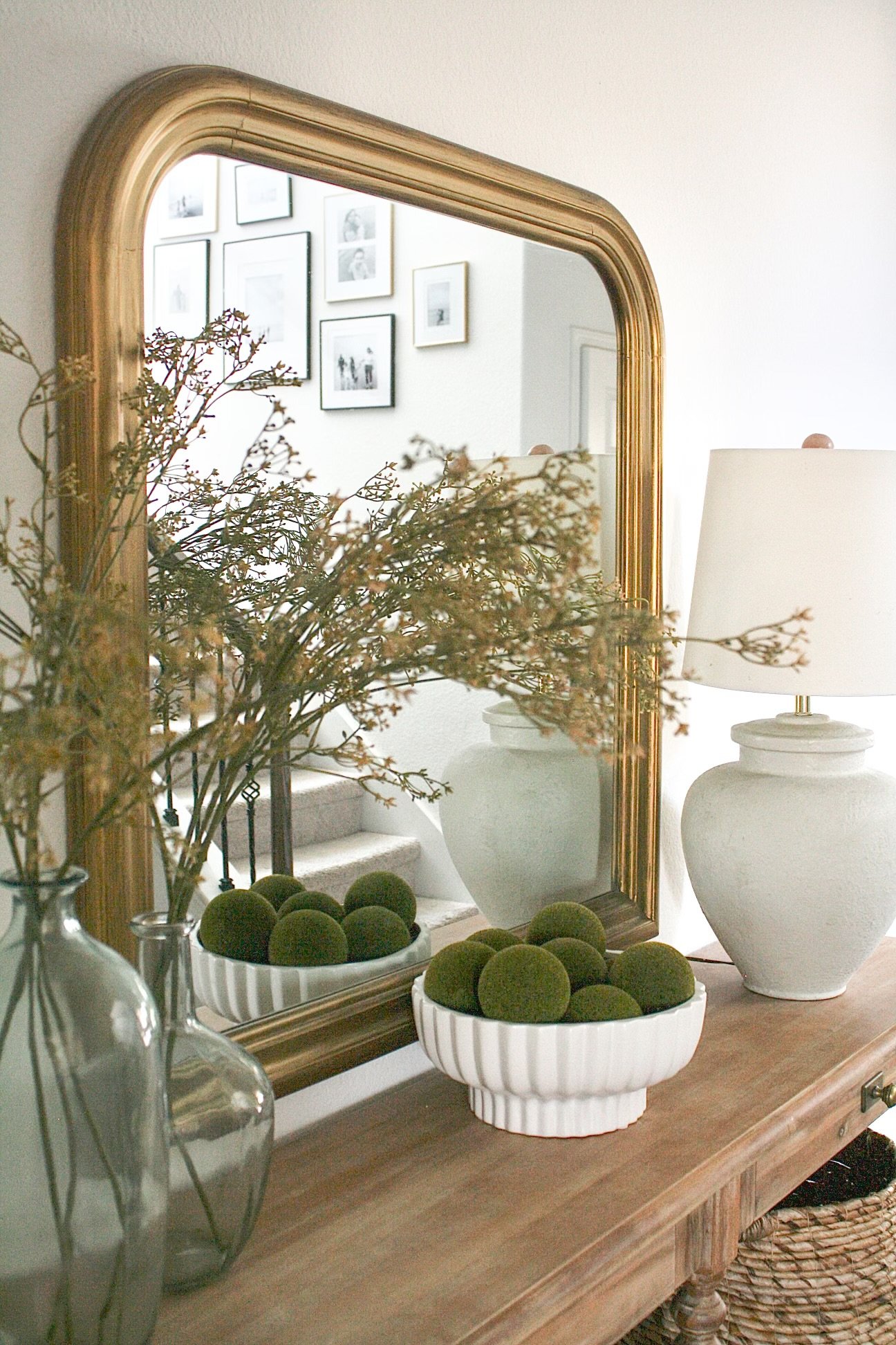 A decorative wooden table with a large mirror, dried plants in a glass vase, moss balls in a white bowl, and a white ceramic lamp with a beige shade. The mirror reflects a white wall with a framed photo gallery. A woven basket is visible at the botto