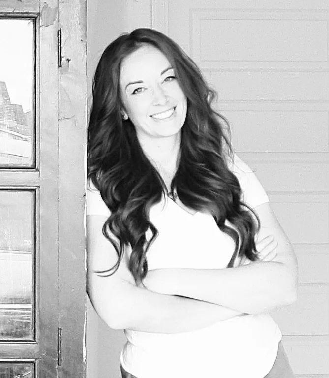 Black and white photo of a woman with long, wavy hair smiling and crossing her arms, standing next to a door.