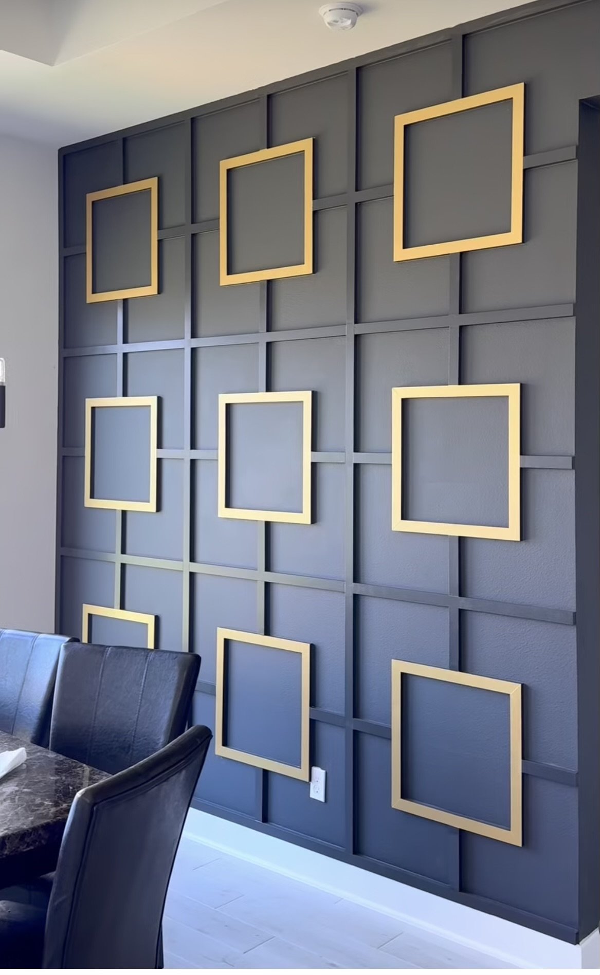 A wall with a grid pattern featuring nine empty, gold collage frames arranged in a 3x3 grid, with a black leather chair and a marble table in the foreground.