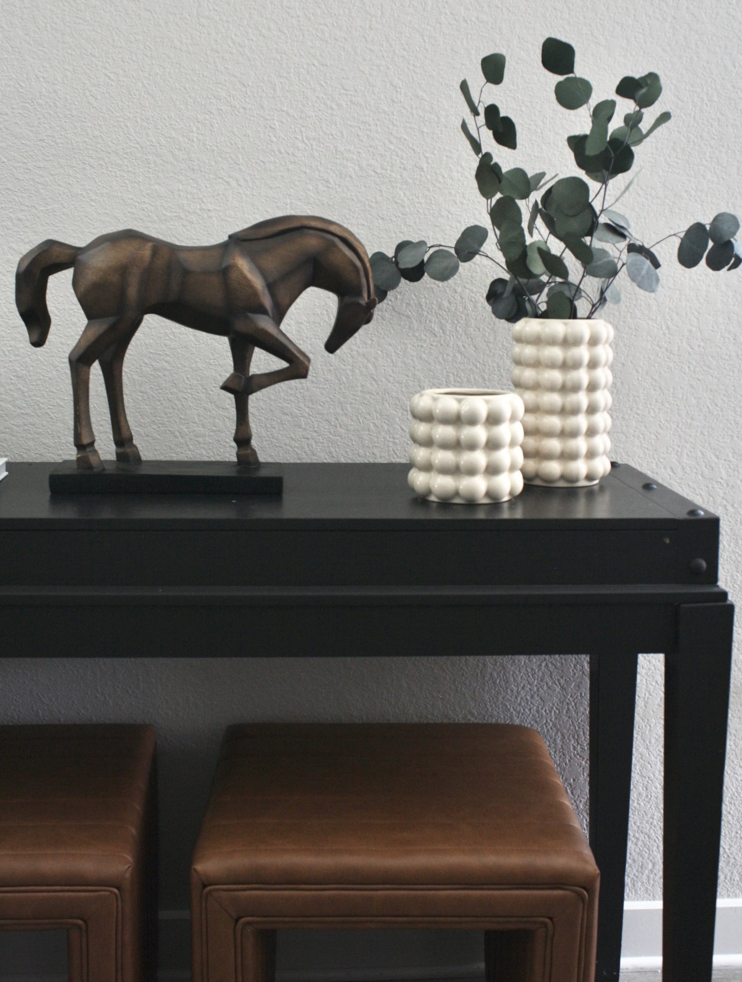 Decorative black console table with a wooden horse sculpture, two white textured vases with green eucalyptus plant, and brown cushioned stools underneath.