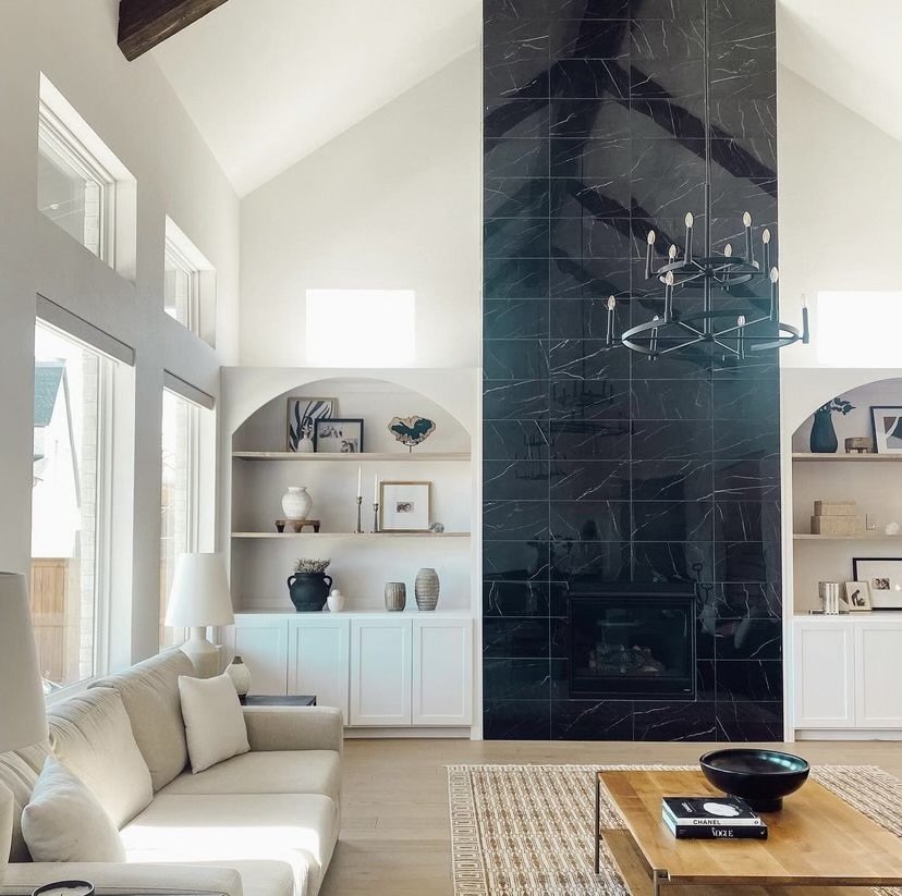 Living room with high vaulted ceiling, large windows, white built-in shelves with decor, a beige sofa, a wooden coffee table with books and a black bowl, and a dark marble fireplace wall with a modern black chandelier.