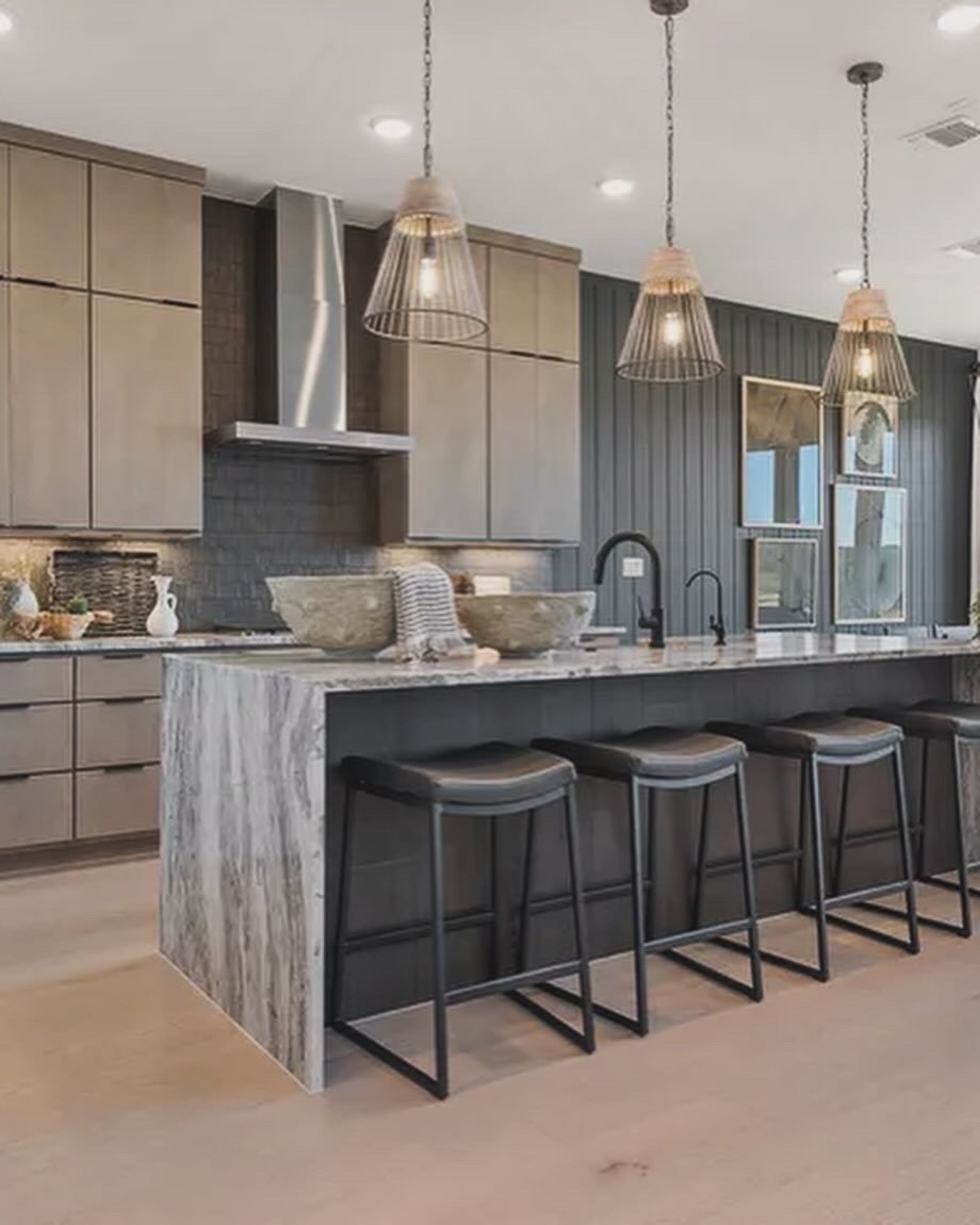 Modern kitchen with a marble island, three black barstools, pendant lights, dark cabinetry, and a window with blinds.