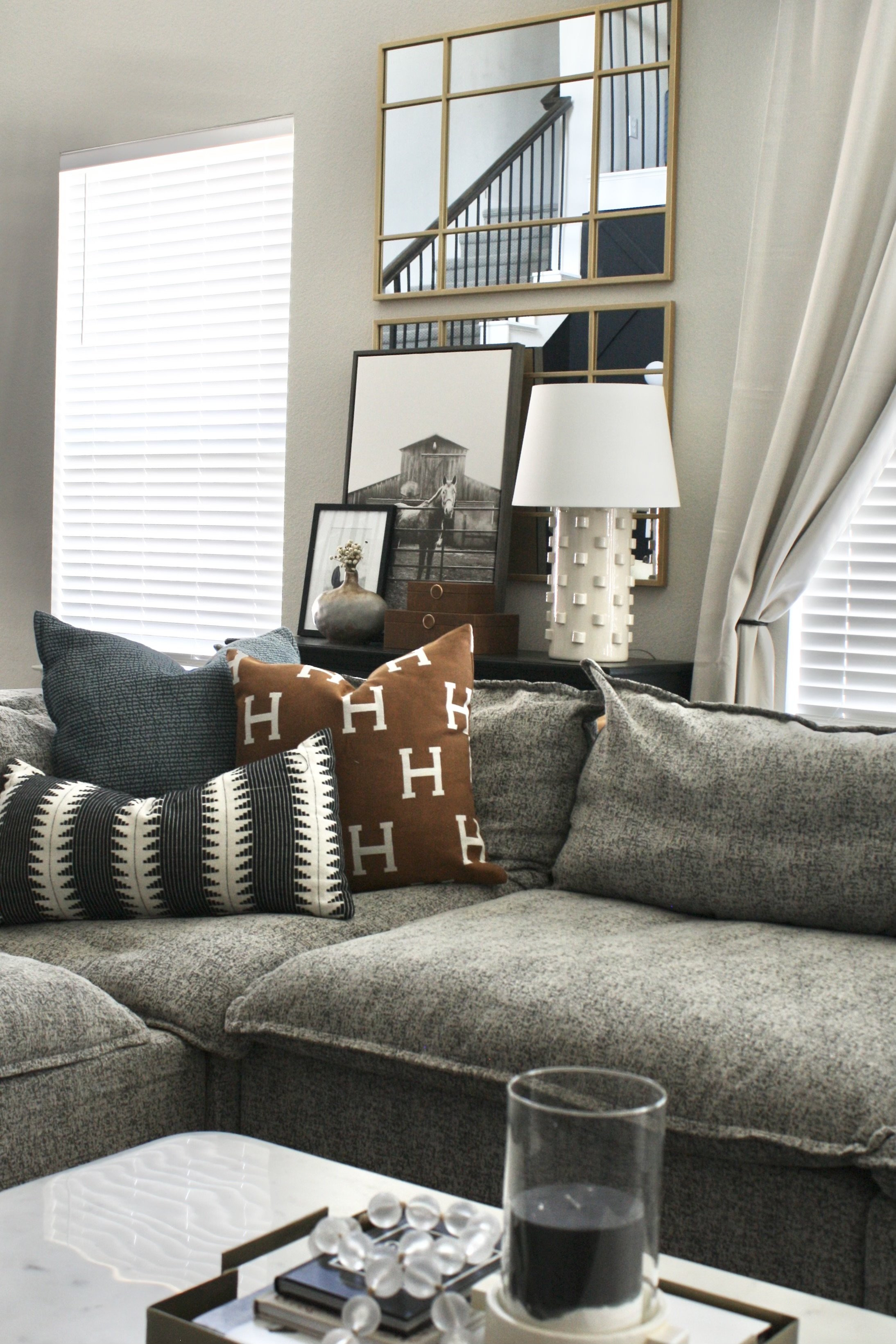 A cozy living room corner with a gray sectional sofa adorned with decorative pillows, a white tray with a glass of dark beverage and a dish with decorative stones or beads, a black side table with framed photographs and a white textured table lamp, a