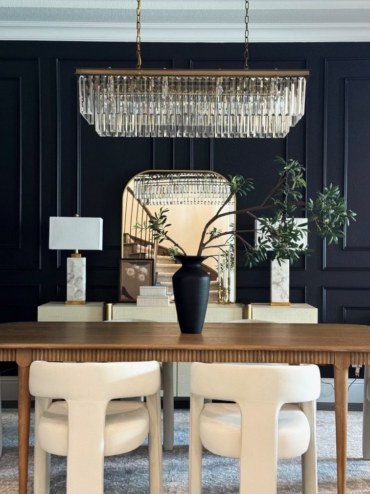 Dining room with a wooden table, two beige chairs, a black vase with green branches, a mirrored wall, and modern lighting fixtures.
