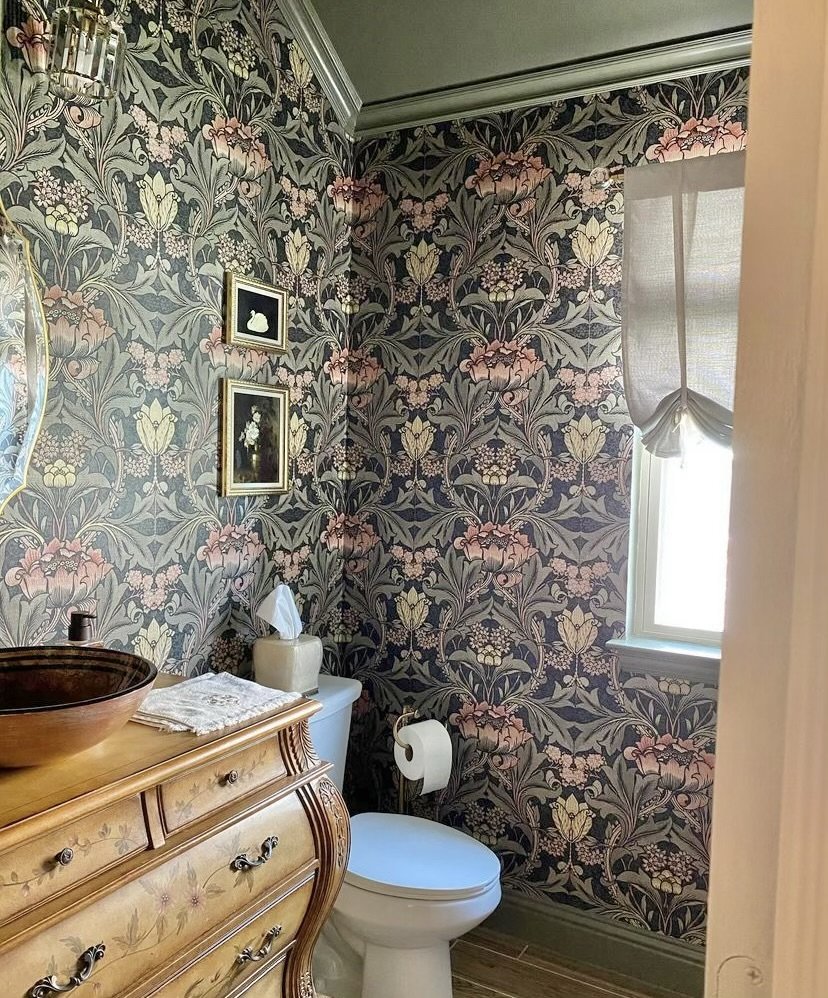 A small vintage-style bathroom with floral wallpaper, a wooden dresser with a vessel sink, a toilet with a tissue roll, a window with sheer curtains, and framed artwork on the wall.