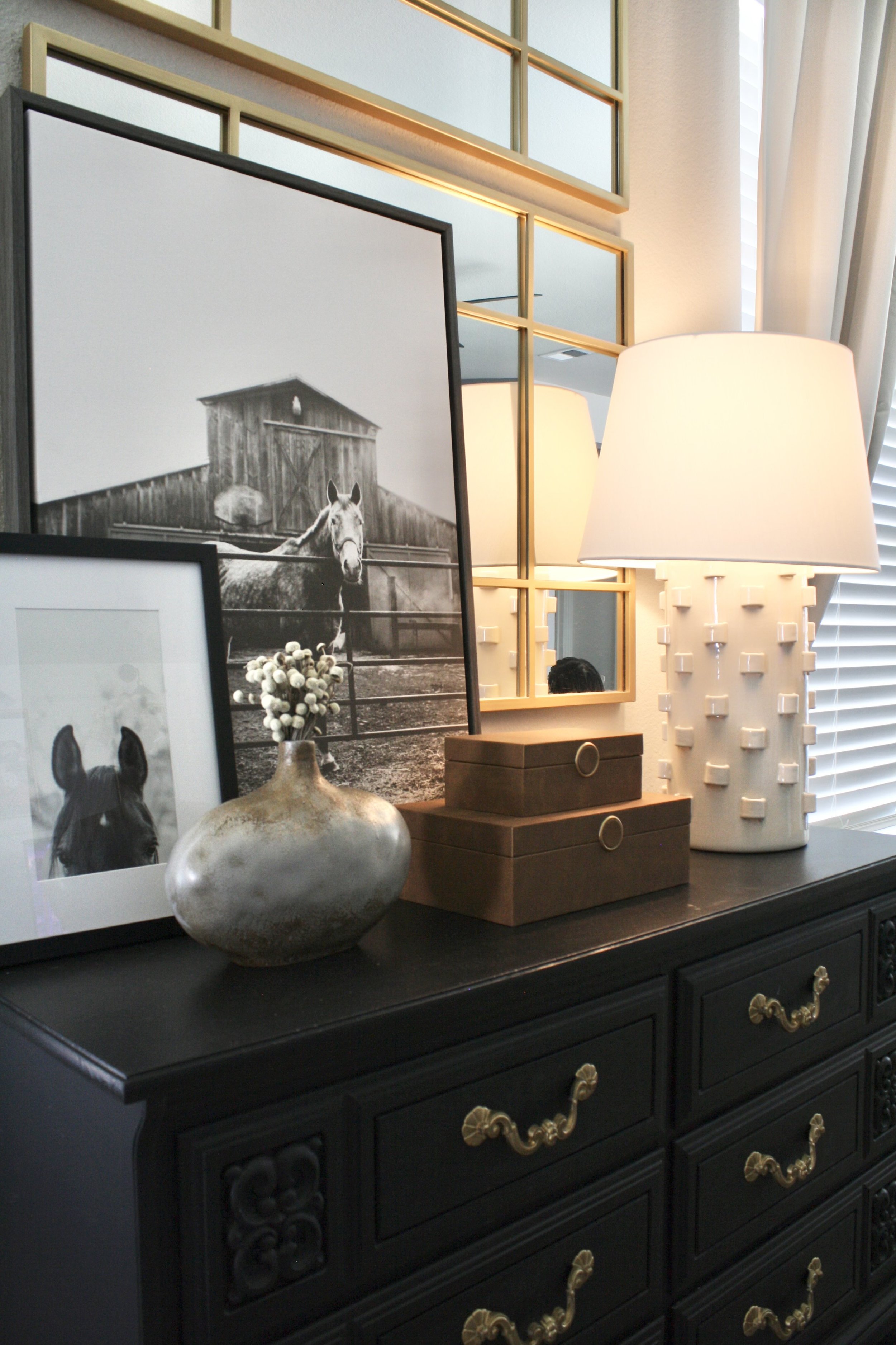 A black dresser with brass handles, decorated with framed horse photographs, a ceramic vase with white flowers, two stacked brown boxes, and a table lamp with a textured white base and a cream shade, in front of a mirror and window blinds.