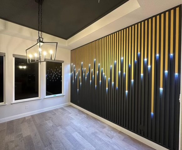 Modern dining room with dark accent wall decorated with vertical black and gold panels and LED light installation, large windows, light wood flooring, and contemporary chandelier.