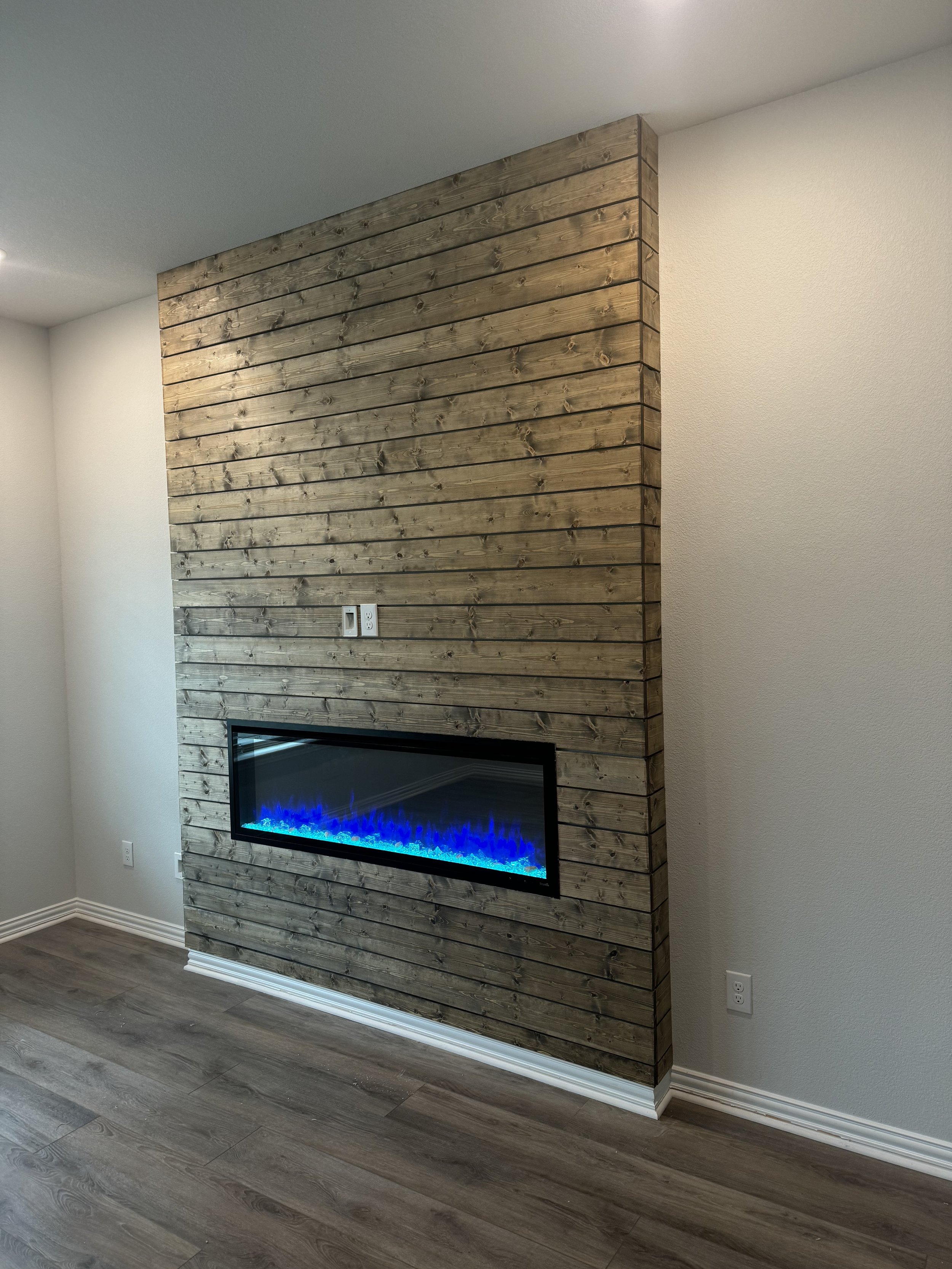 Interior of a room featuring a modern built-in electric fireplace with blue flames, surrounded by a wall of horizontal wooden planks, adjacent to white walls and floors with baseboards.