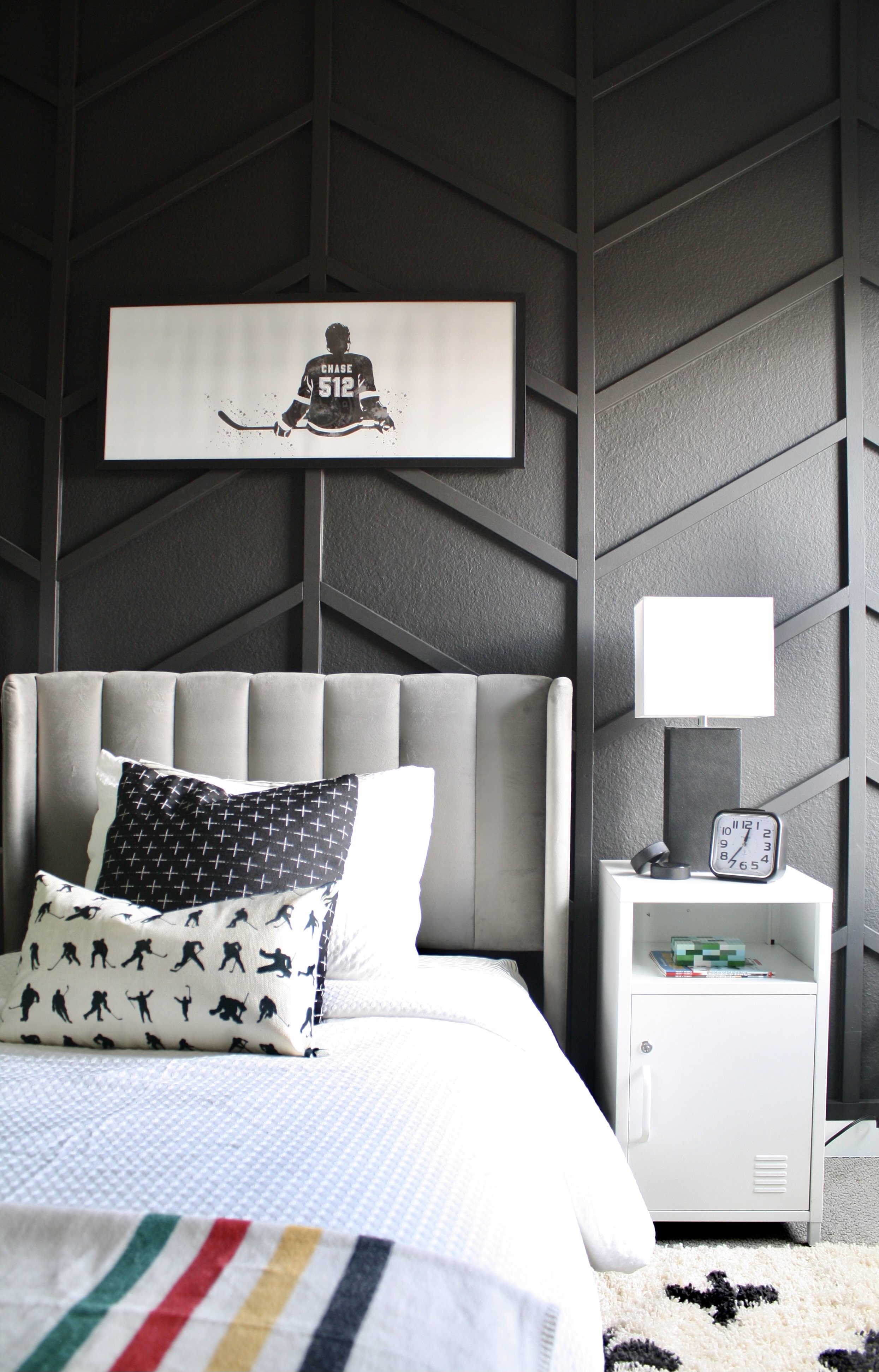 Bedroom with a beige upholstered headboard, decorative pillows featuring hockey player silhouettes, a white nightstand with a white lamp, a black alarm clock, a telephone, and books. A framed hockey player silhouette photo hangs on a textured black w