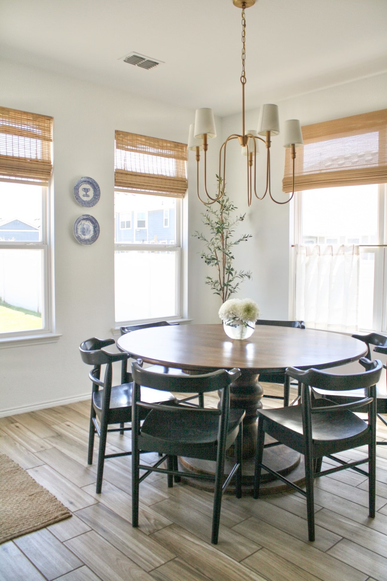 Dining room with round wooden table, six black chairs, a hanging chandelier, three windows with bamboo shades, a potted plant, and a ceramic flower arrangement on the table.