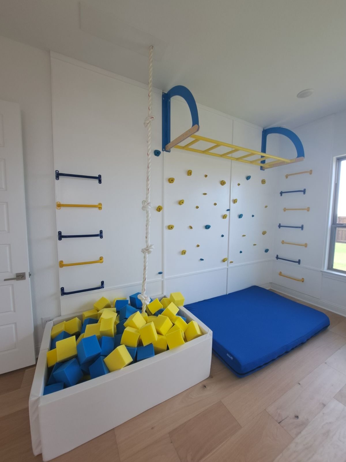 Indoor children's playroom with climbing wall, foam pit filled with yellow and blue foam blocks, gym mat, and climbing ladder.