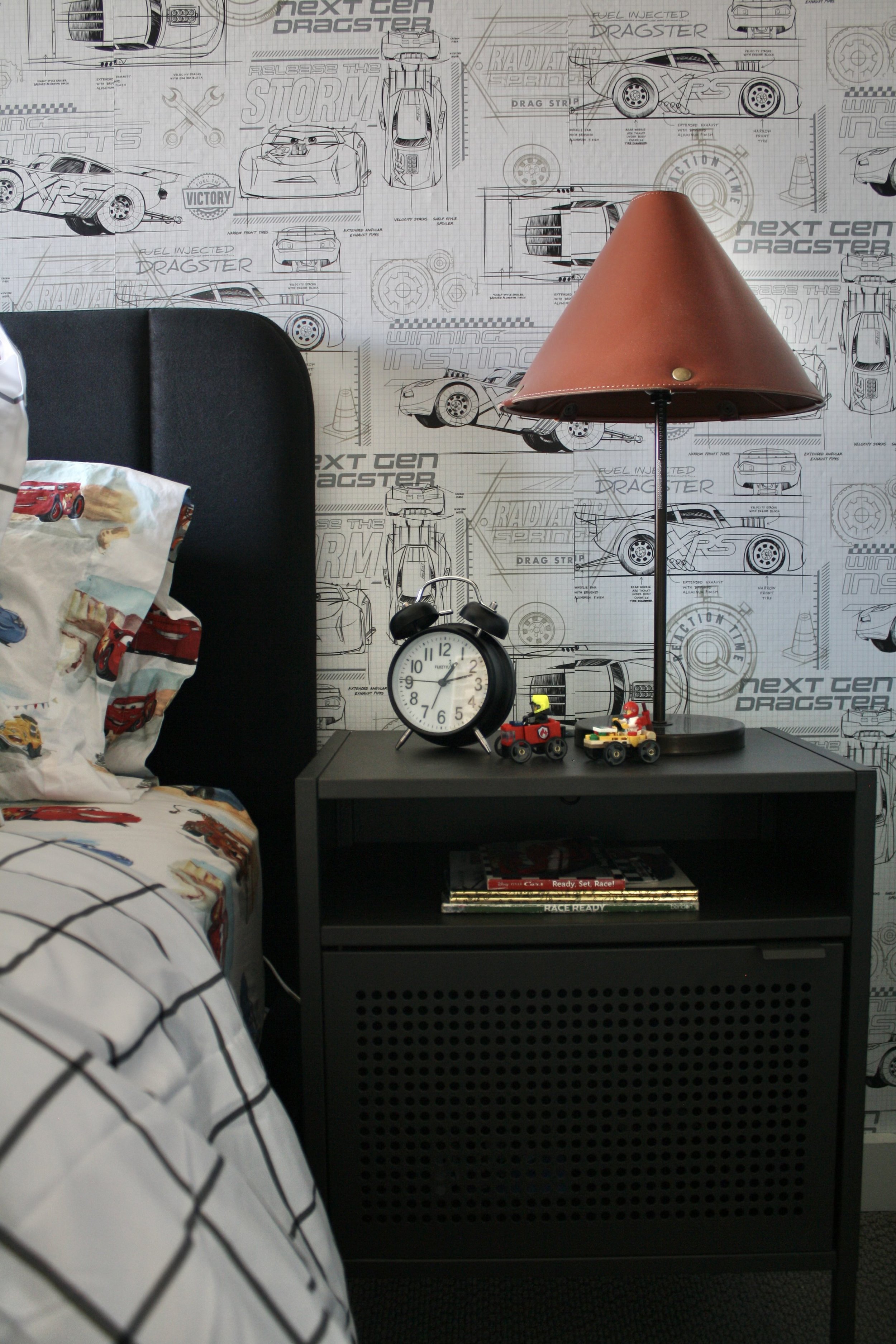 A modern black nightstand with a vintage alarm clock, Lego cars, and a table lamp with a brown shade. Behind the nightstand is a wallpaper with car blueprints and racing themes. Part of a bed with white sheets and pillow featuring car graphics is vis