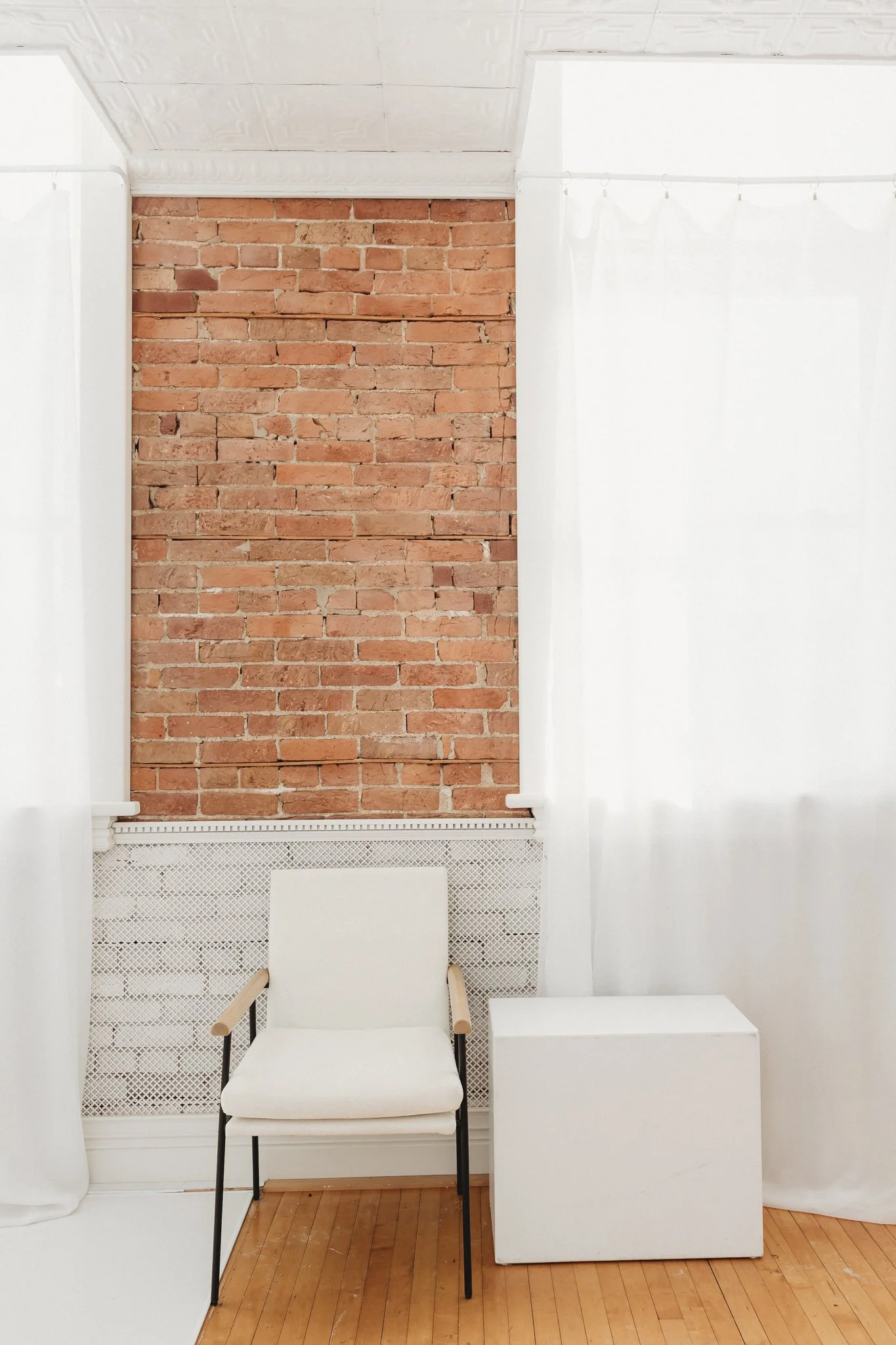 Empty white chair with wooden armrests, white cube table, and brick wall behind, within a room with white curtains and hardwood floor.