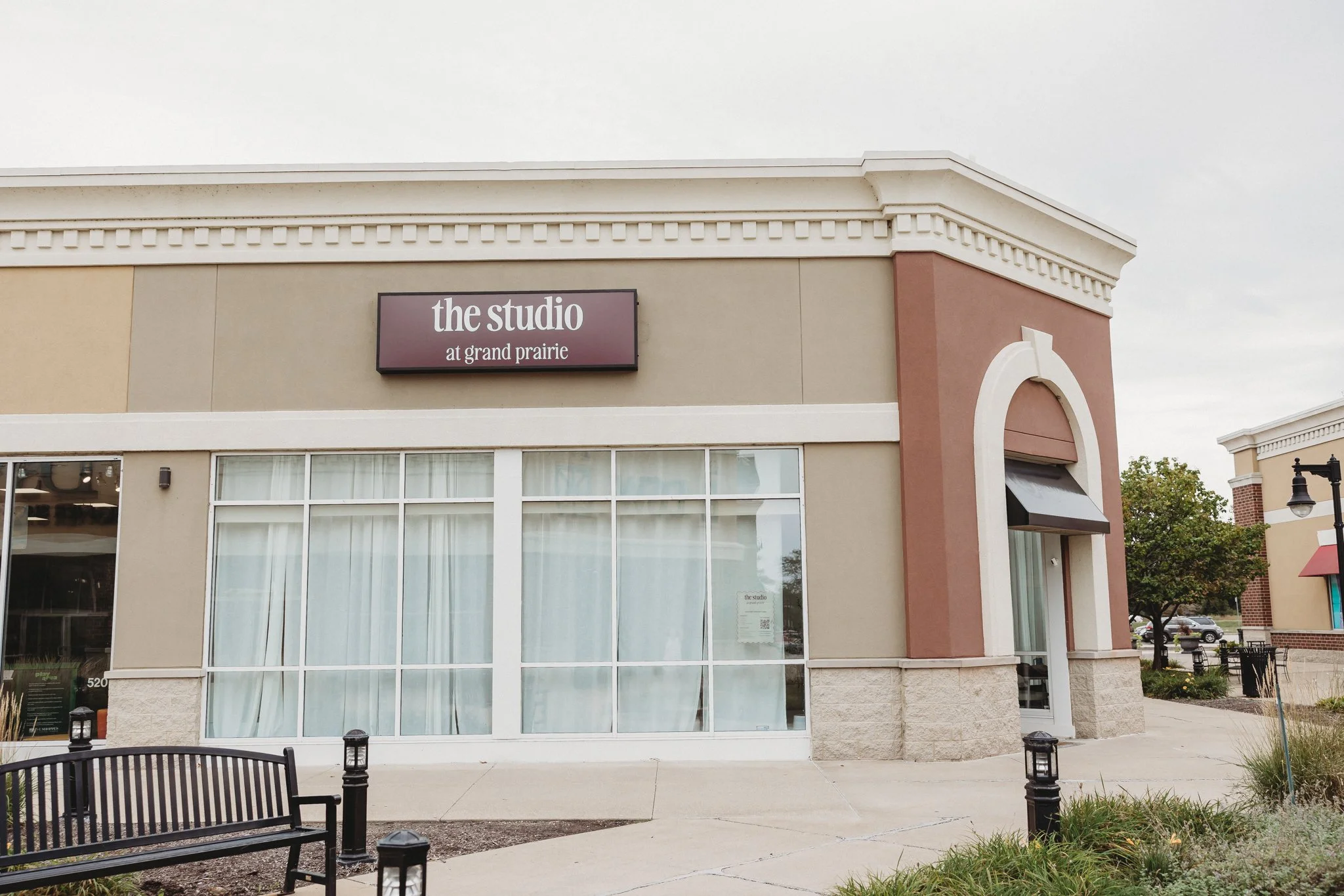The exterior of a commercial building with a sign that reads "the studio at grand prairie." The building has large windows, a beige and pink facade, and decorative architectural details. There are benches, small lantern-style lights, and trees in the surrounding area.