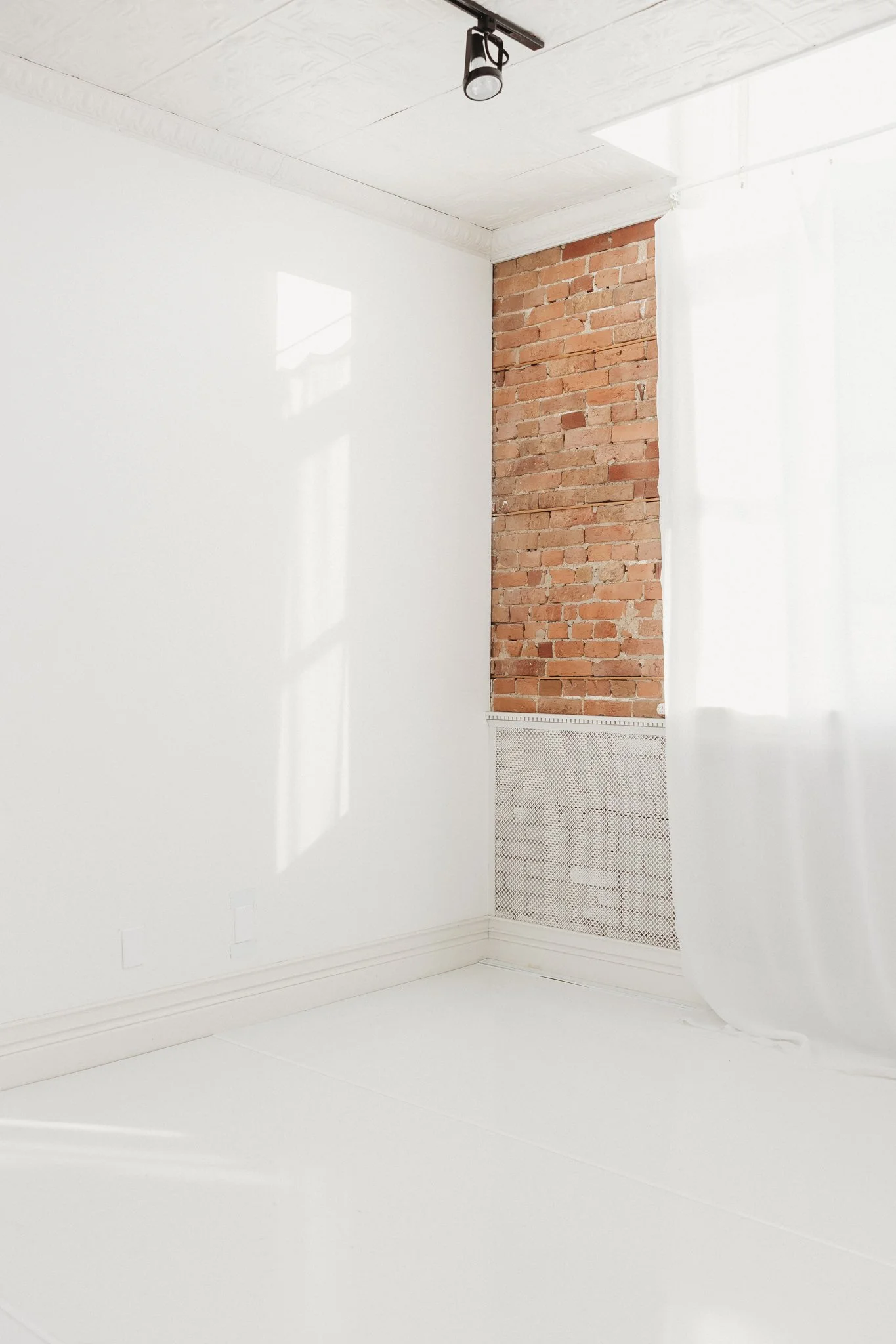 An empty room with white walls, a brick wall section, a white curtain, a ceiling light fixture, and sunlight casting shadows on the wall.