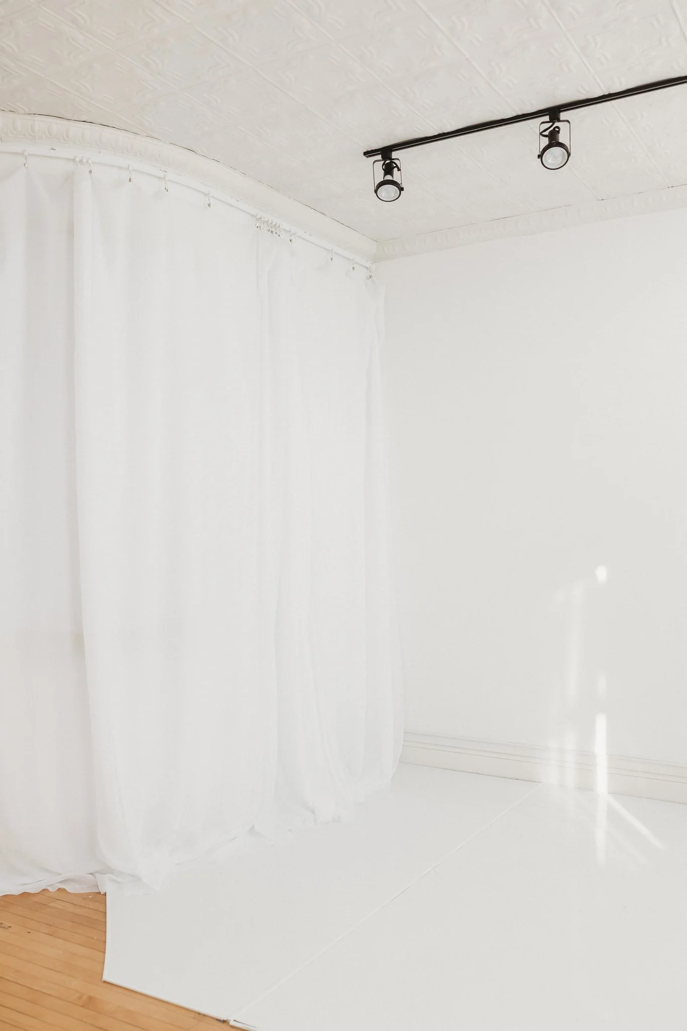 Empty room with white walls, white curtains, wooden floor corner, and ceiling with track lighting.