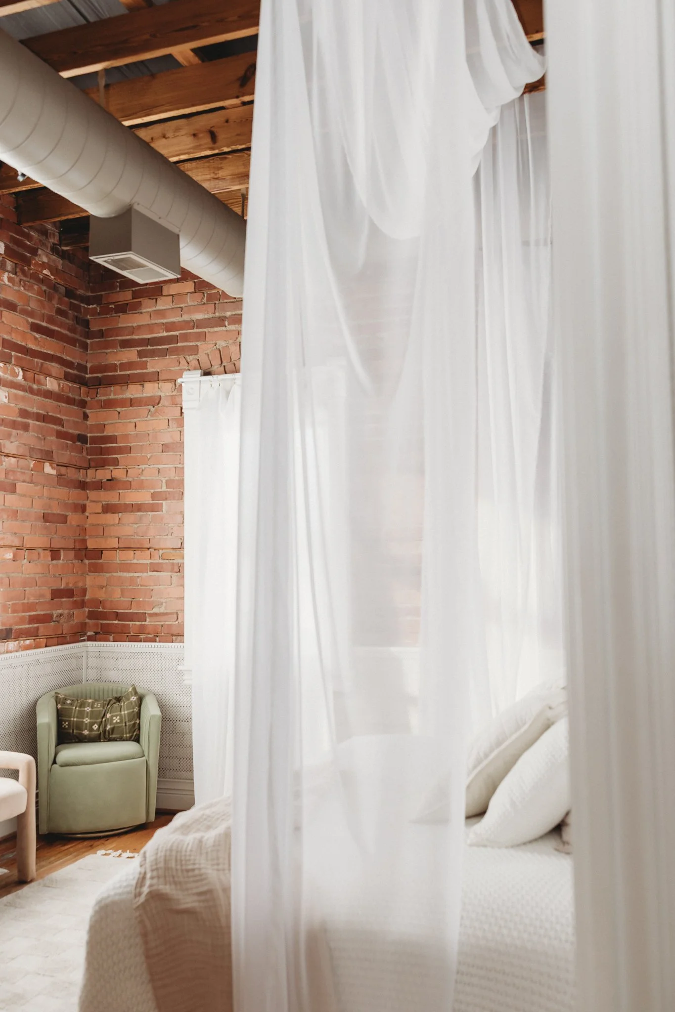 A cozy bedroom with a white canopy bed, sheer white curtains, and a brick wall. There's a light green armchair with a decorative pillow and part of a beige armchair visible. The ceiling has exposed wooden beams and ductwork.