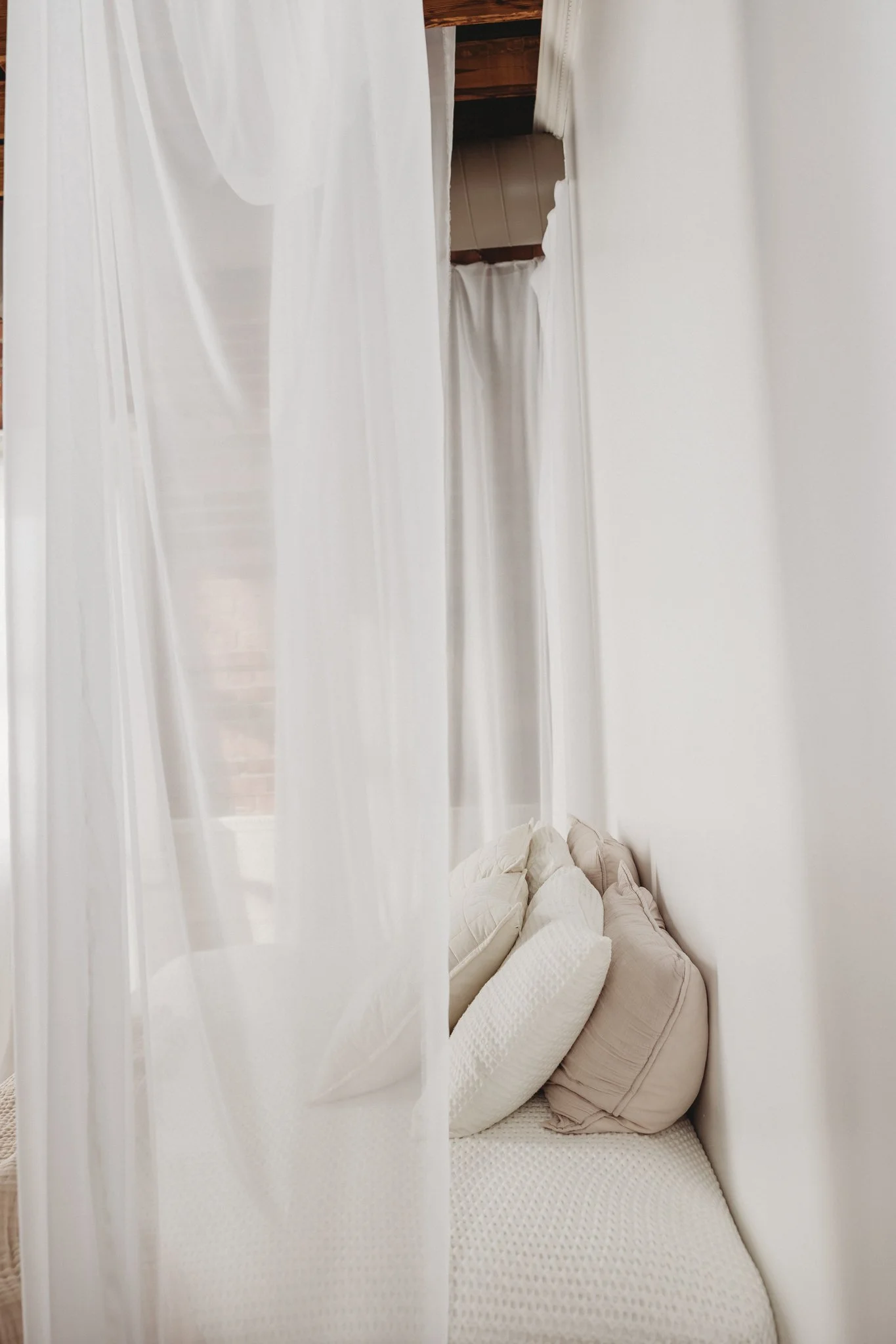 A cozy corner with a bed covered in white textured bedding and an arrangement of white and beige pillows. White curtains surround the space, with a wooden ceiling visible above.