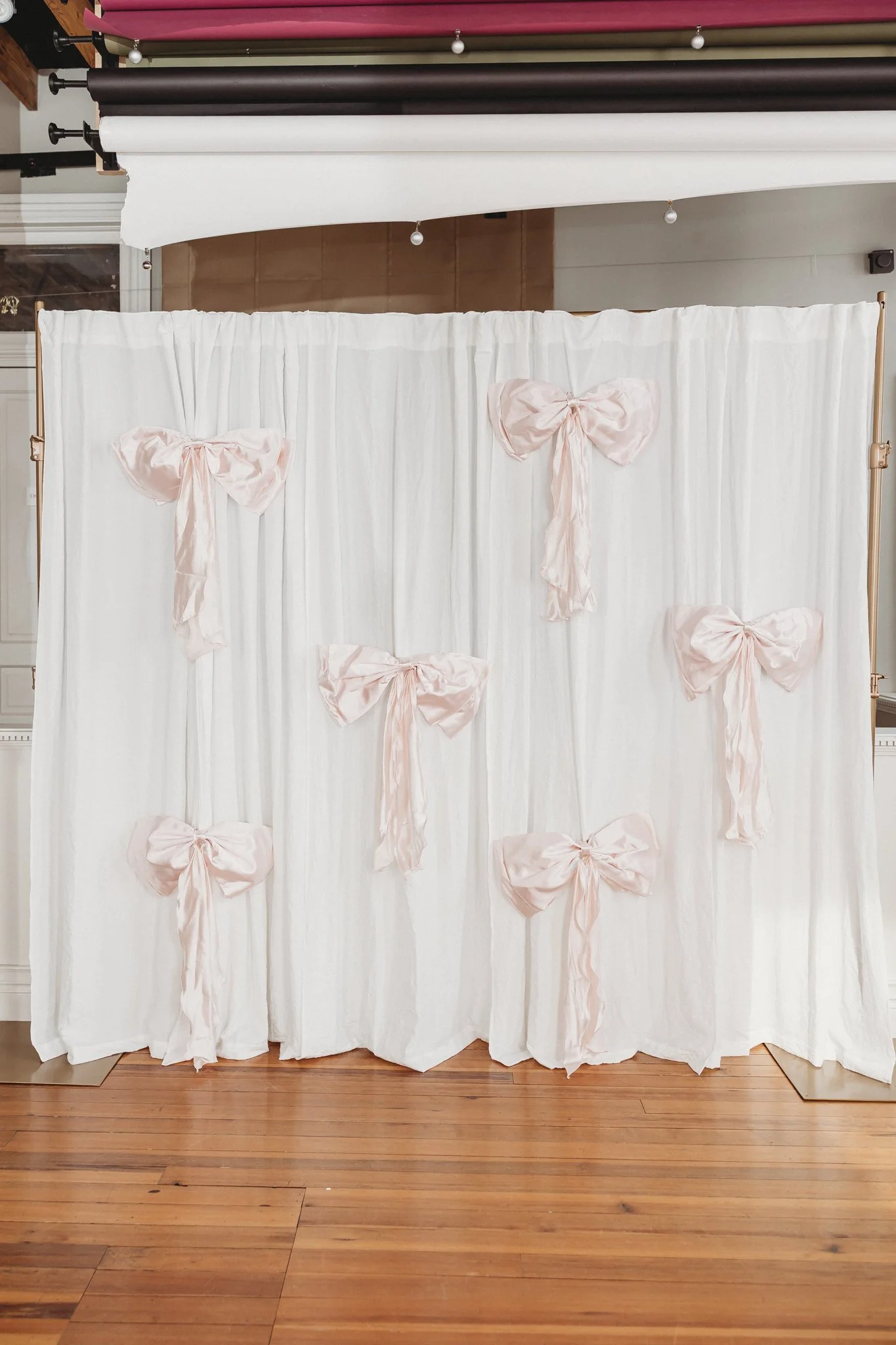 White fabric backdrop decorated with pink satin bows, arranged in a grid pattern, on a wooden floor in a room.