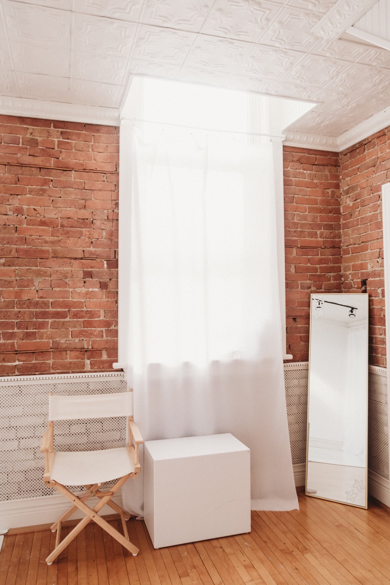 Interior room inside a rentable photography studio in Tremont, Illinois, with a brick wall, white curtain, wooden floor, standing mirror, small wooden chair, and white cube.