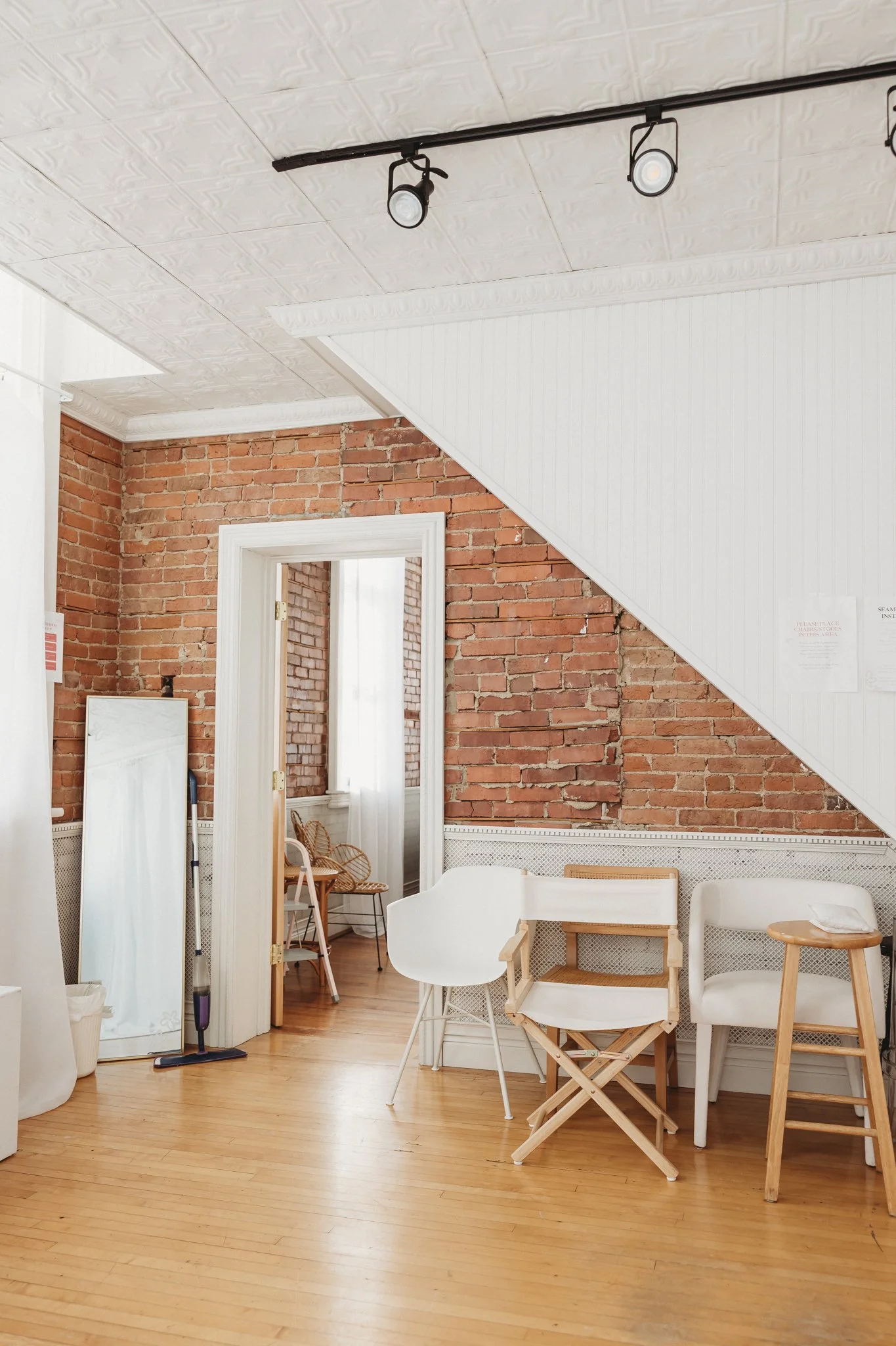 Interior of a room featuring exposed brick walls, white furniture, wooden flooring, and an angled white ceiling with track lighting.