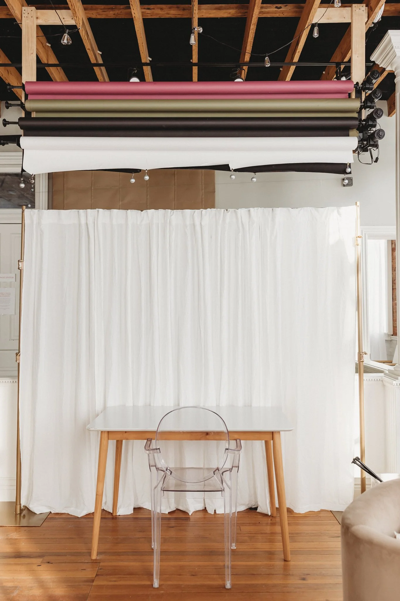 Photograph of a photography studio setup with a white curtain backdrop, a wooden table, a transparent chair, and color prints of fabric rolls hanging from pipes on the ceiling.