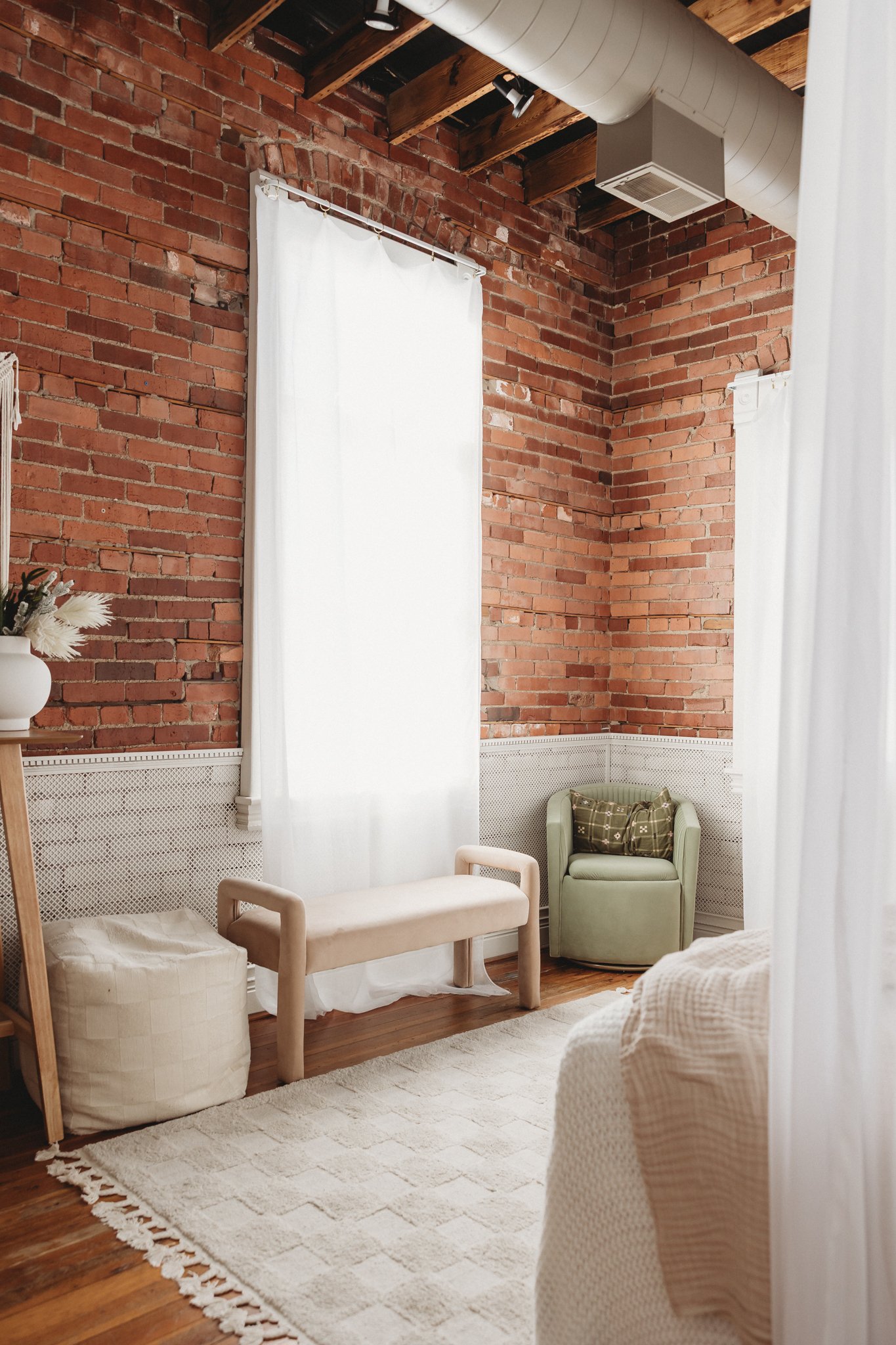 A cozy corner of a room with exposed red brick walls, white curtains, a beige bench, a green armchair with a patterned pillow, a white textured ottoman, and a cream-colored rug.
