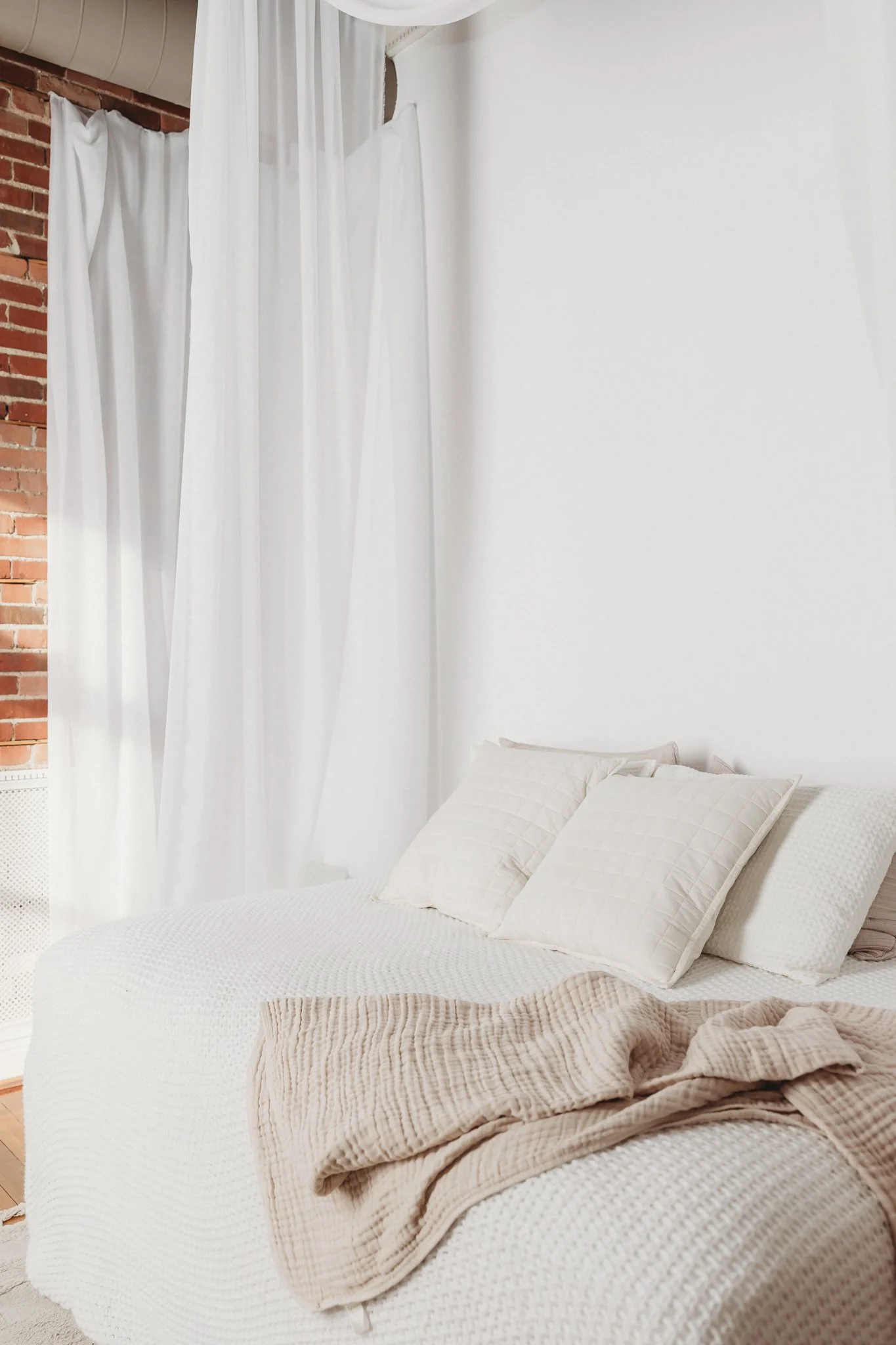 A bedroom with white bedding, two pillows, a beige blanket, white curtains, and part of a brick wall.